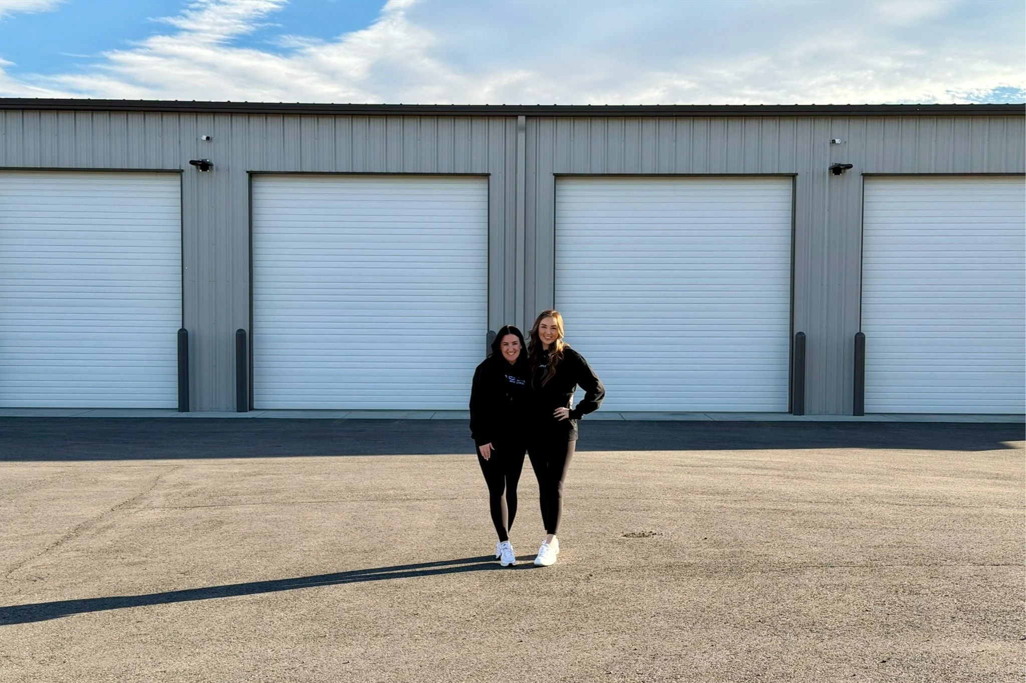 Two women standing in front of a large industrial building with three closed garage doors.