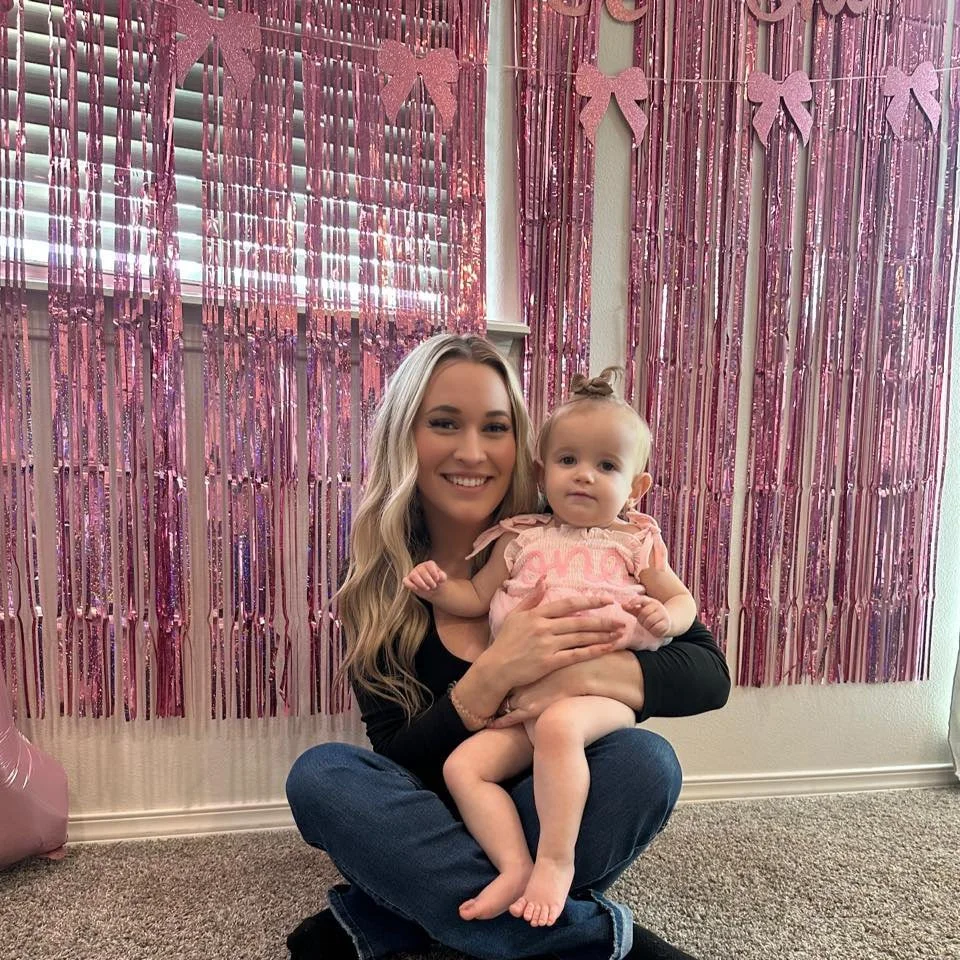 A woman with long blonde hair, sitting cross-legged on a carpeted floor, holding a young girl in front of a pink metallic tinsel backdrop with bows. The woman is smiling and wearing a black top and blue jeans. The girl has light hair in a ponytail, wearing a pink dress, and has a neutral expression.