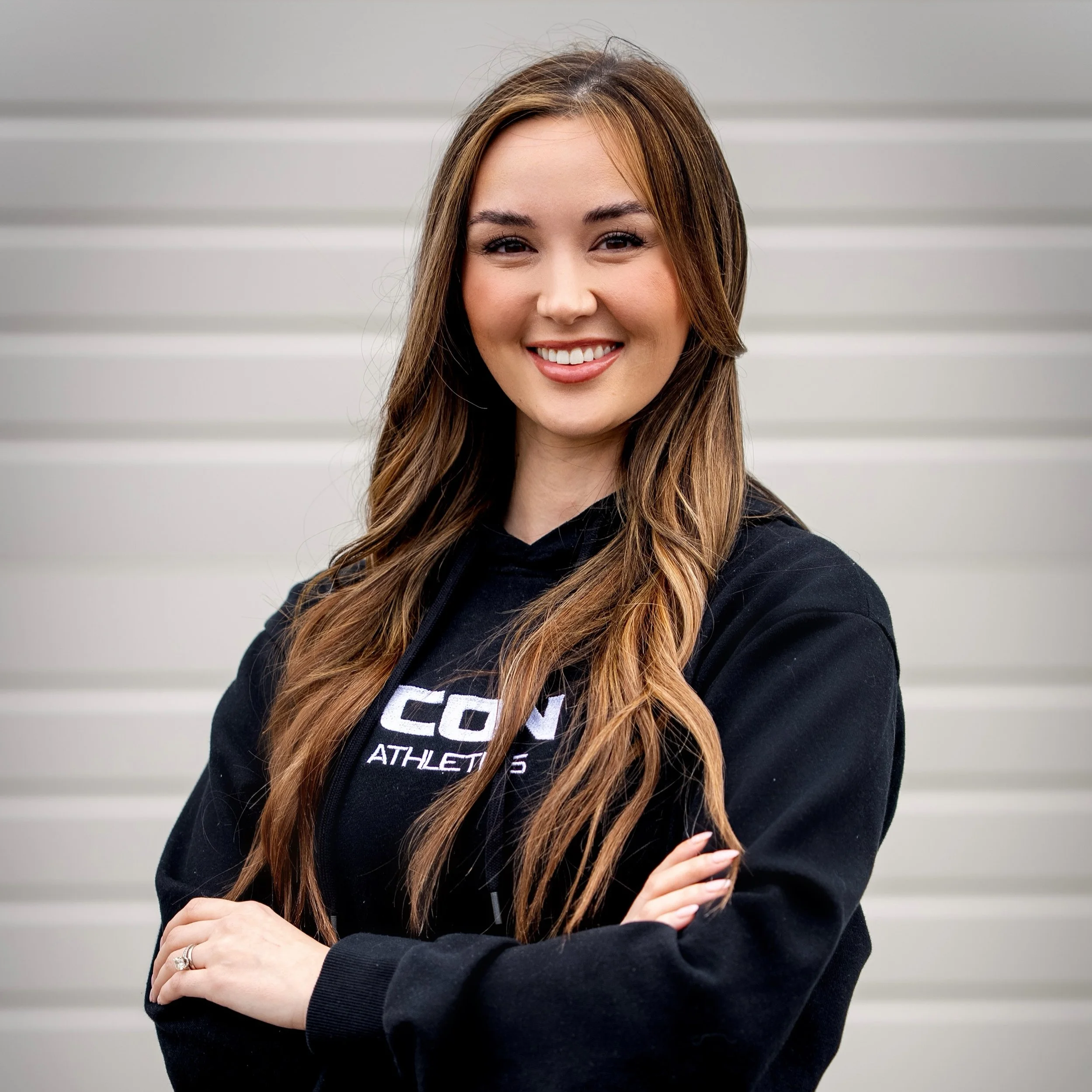 A smiling woman with long brown hair wearing a black hoodie with white text, standing in front of a gray horizontal wall.