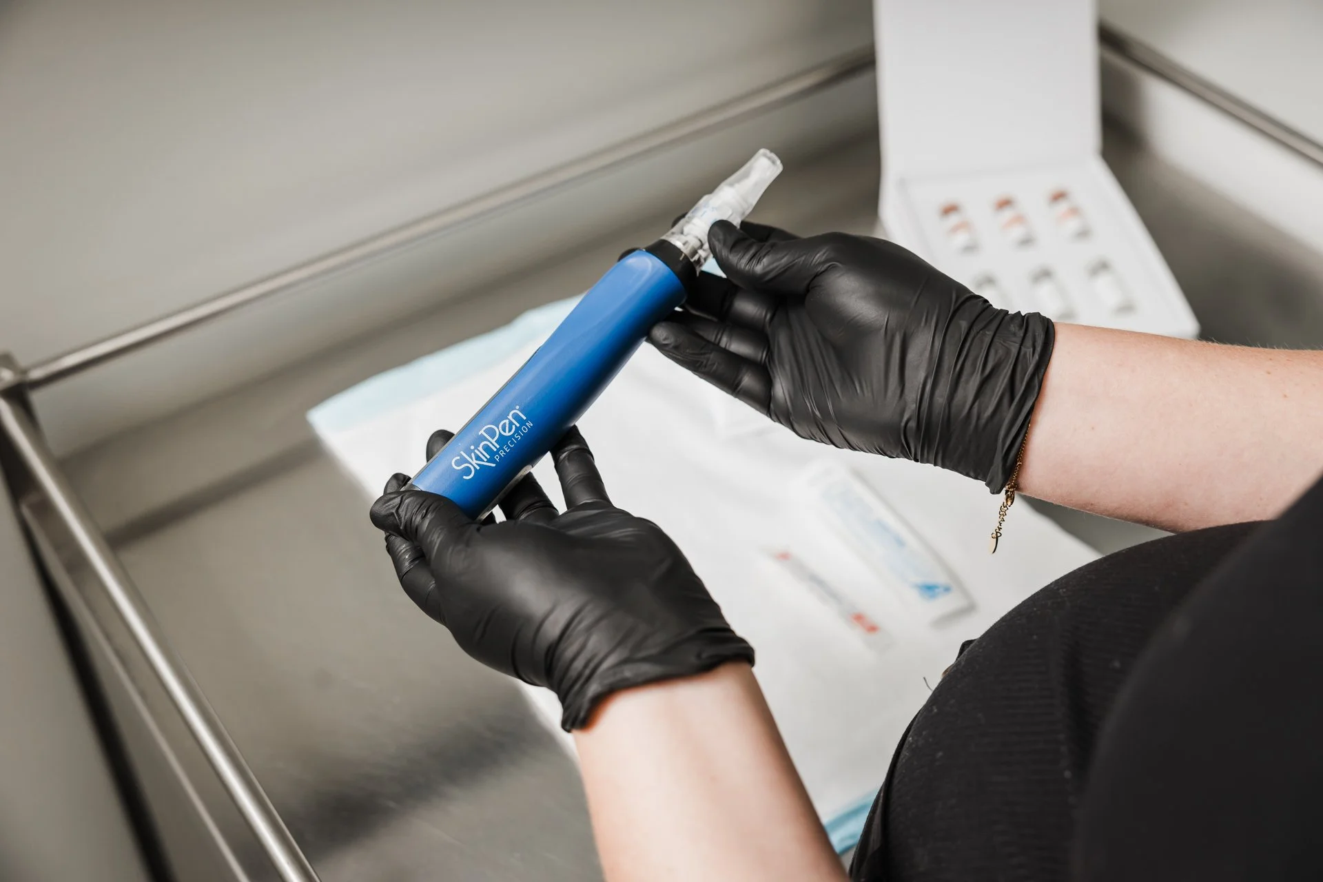 Person wearing black gloves holding a blue SkinPen Precision micro-needling device over a sterile tray with medical supplies.