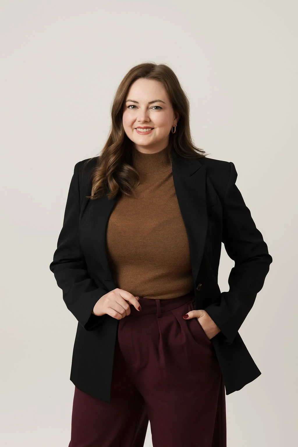 A woman with long brown hair wearing a black blazer, brown sweater, and burgundy pants, standing against a plain light-colored background.