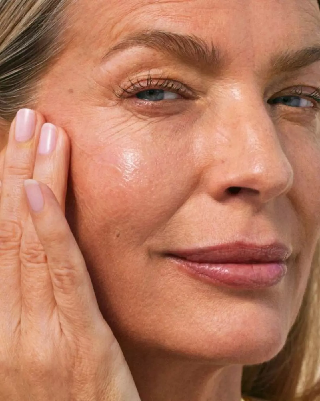 Close-up of a woman touching her cheek with her fingers, showing clear and smooth skin, with visible fine lines and makeup.