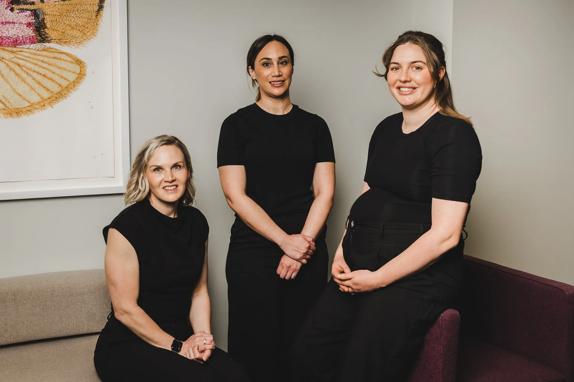 Three women in black clothing posing in an indoor setting with a framed wall art piece on the wall.