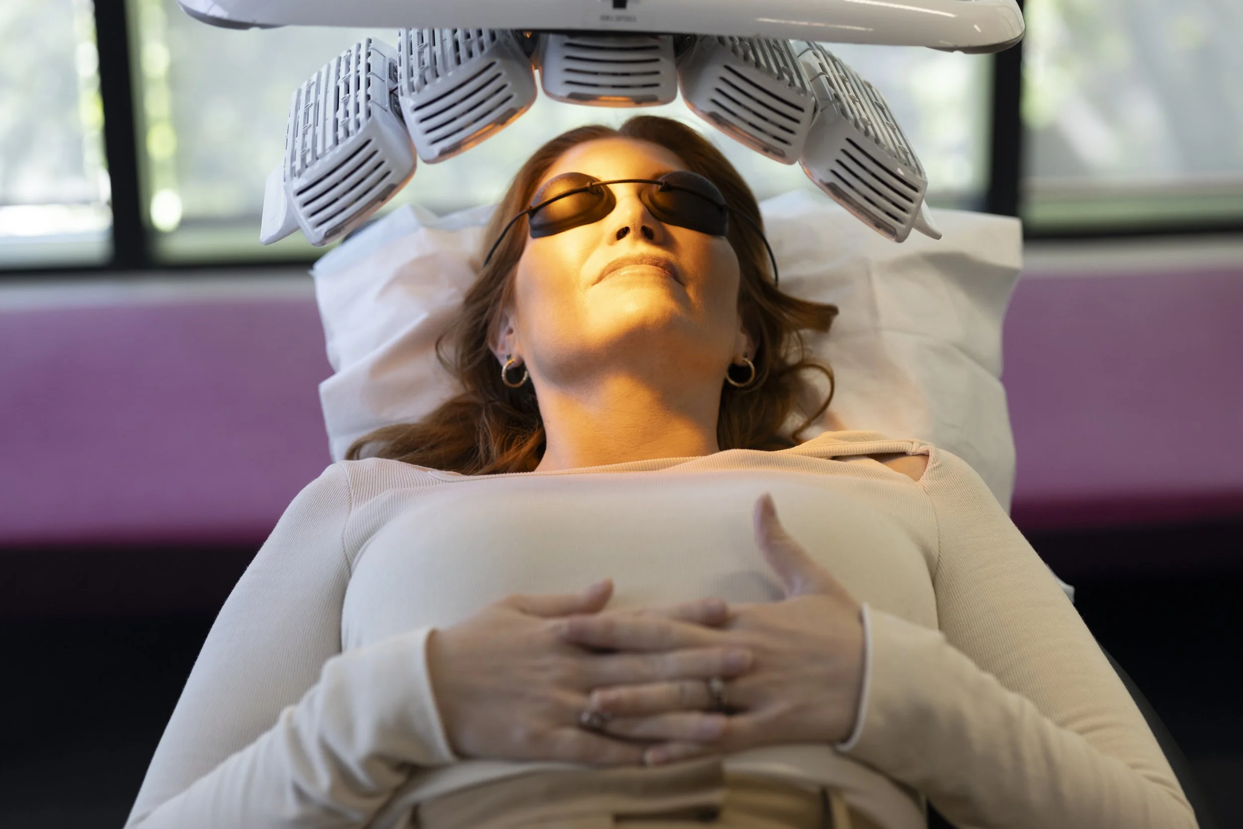 A woman lying on a medical bed with her eyes covered by eye protection, with a heat lamp or medical device positioned above her head, in a clinical setting.