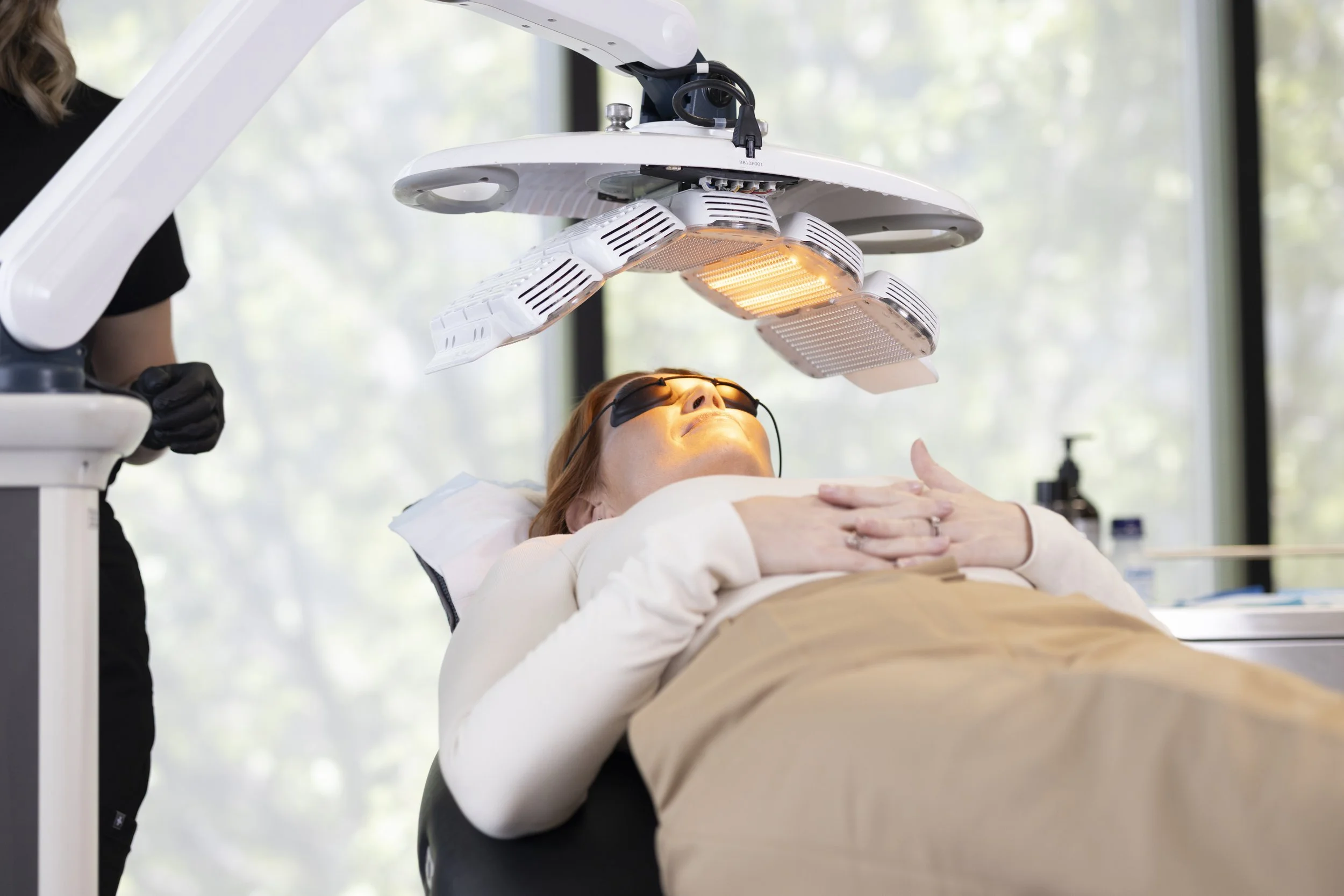 A woman lying on a medical bed undergoing a light therapy treatment with protective glasses, while a medical professional operates a device above her.