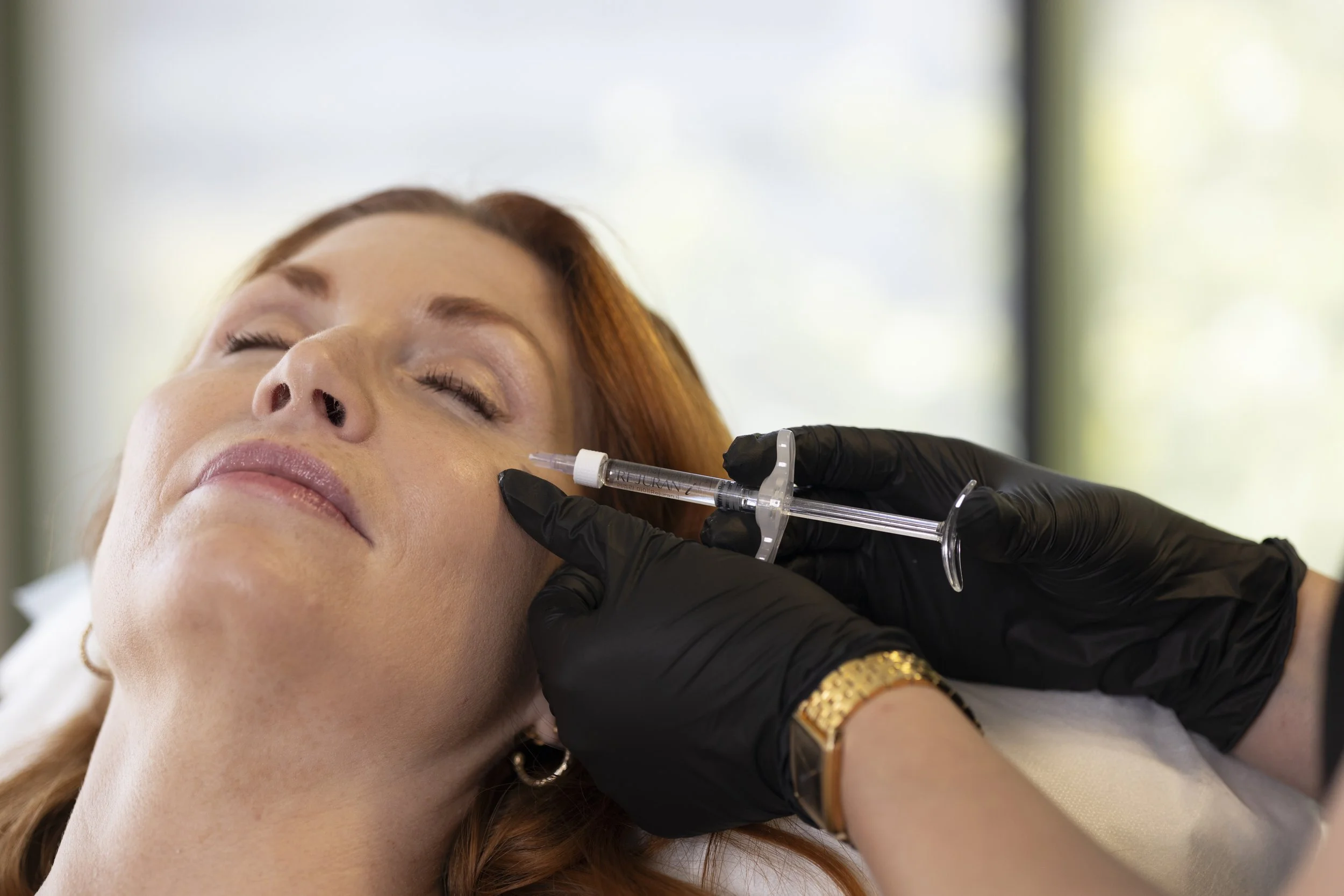 A woman receiving a cosmetic injection in her cheek during a medical procedure, with the healthcare professional wearing black gloves.