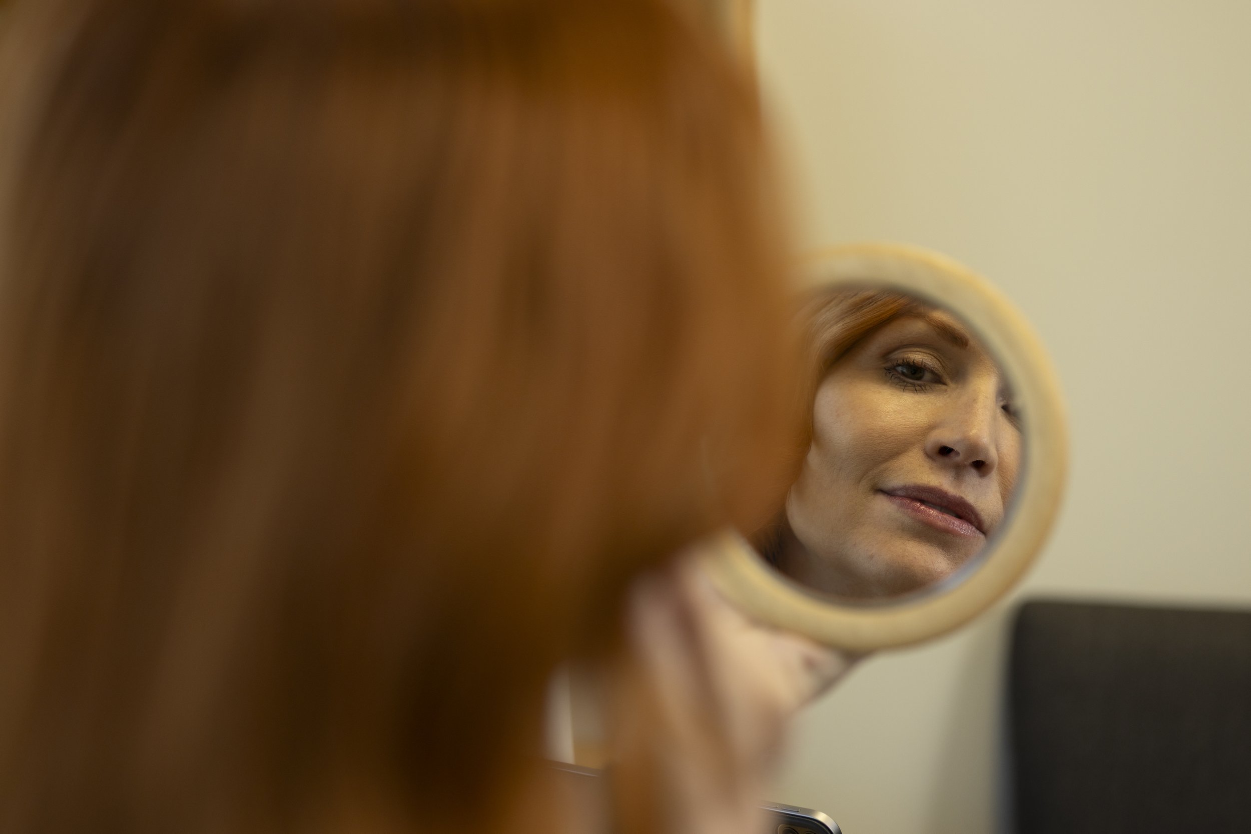 A woman with red hair looking into a small round mirror, her face reflection visible in the mirror.