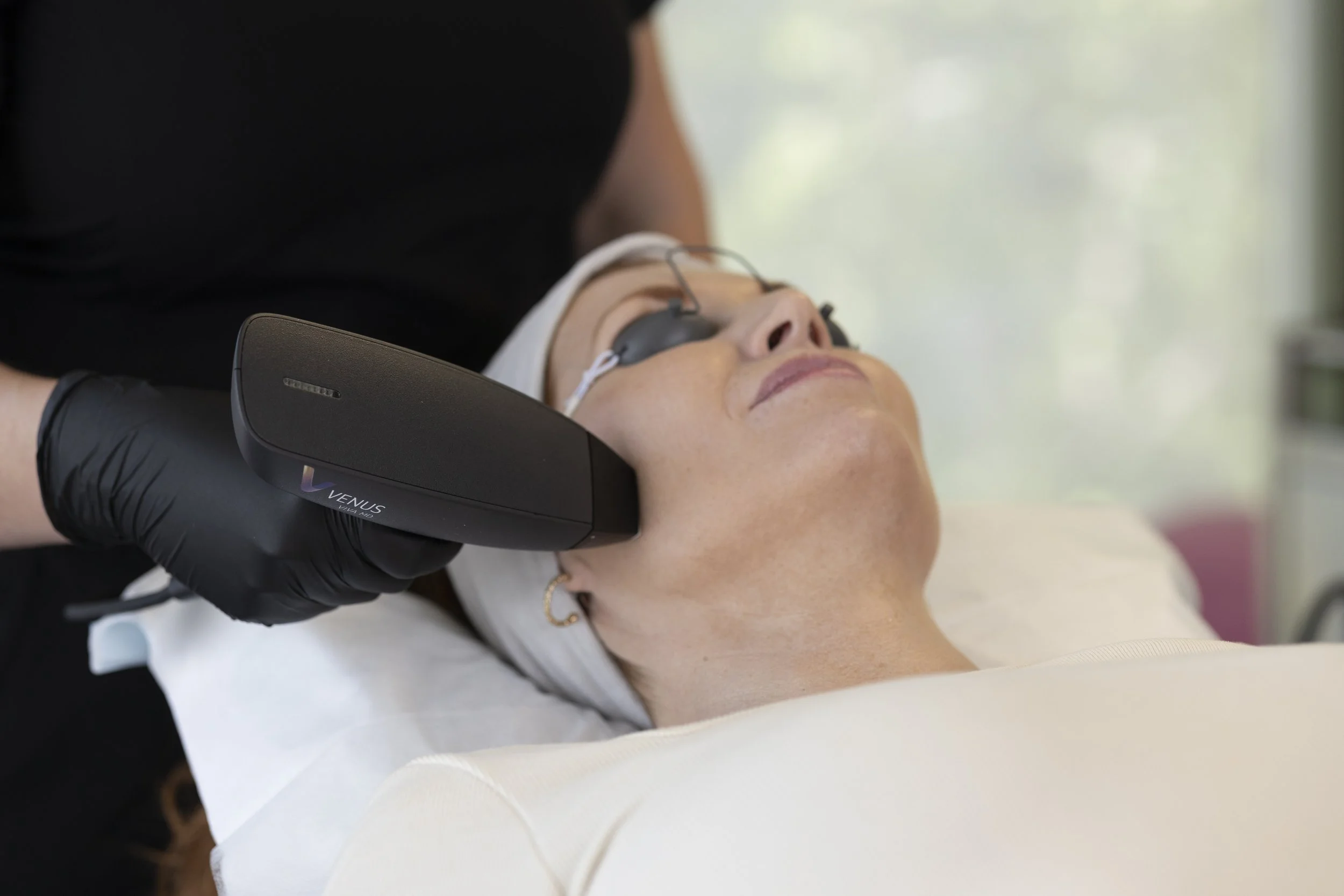 A woman is lying on a treatment bed with her eyes covered, receiving a facial treatment with a Venus machine.