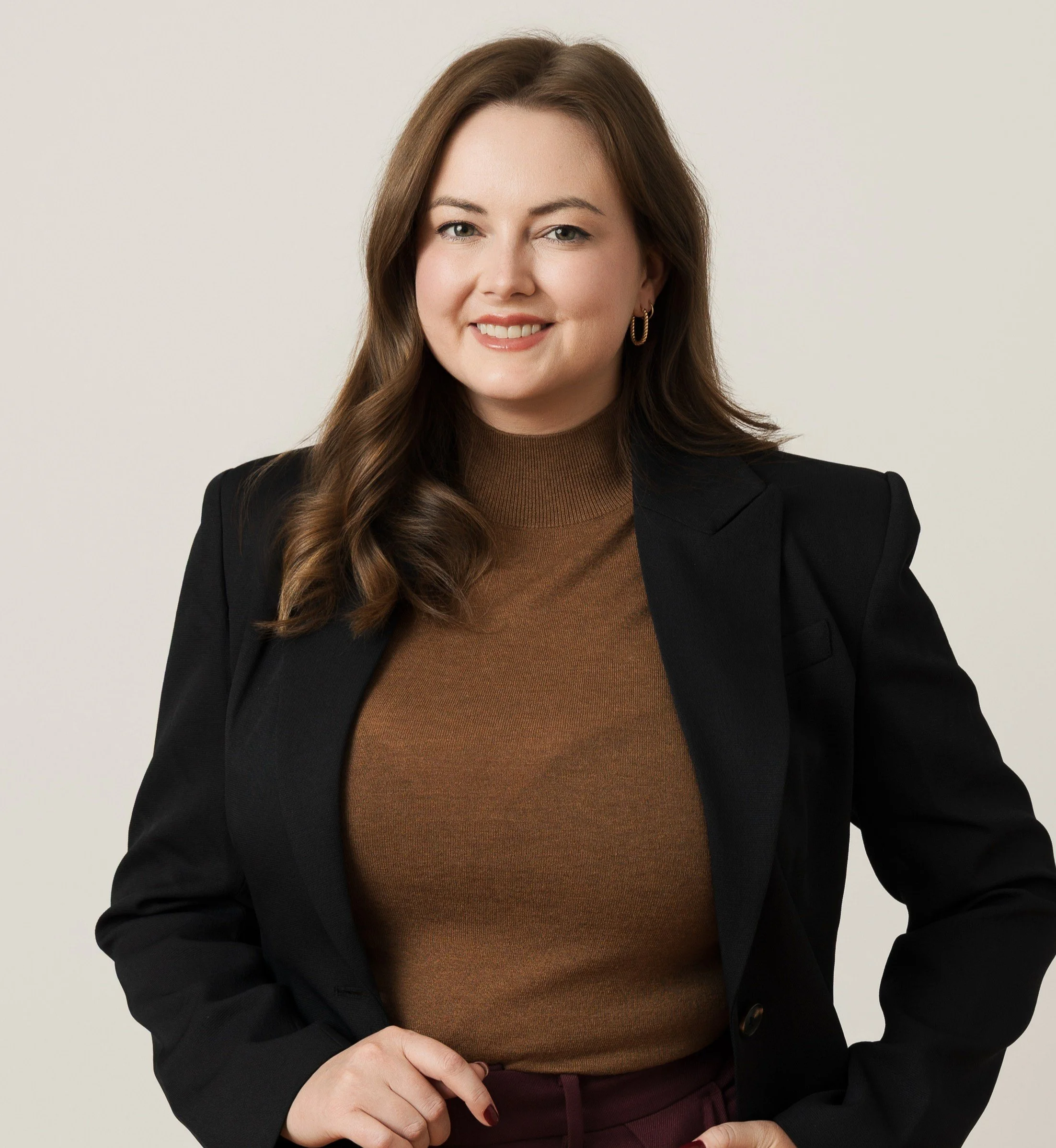 a young female doctor with wavy brown hair, smiling at the canberra wearing a black blazer, brown knitted top and burgundy pants, with a neutral background