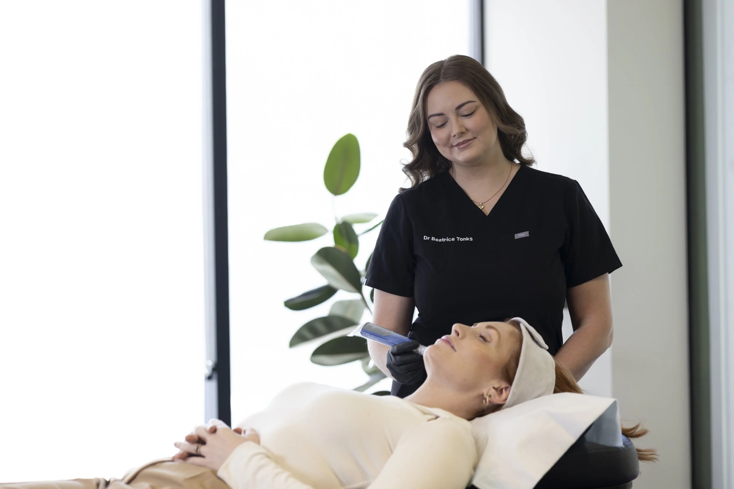 A woman lying on a treatment bed with her eyes closed. A woman dressed in black medical scrubs and gloves is standing over her, holding a small device near her face. There is a large window and a green plant in the background.