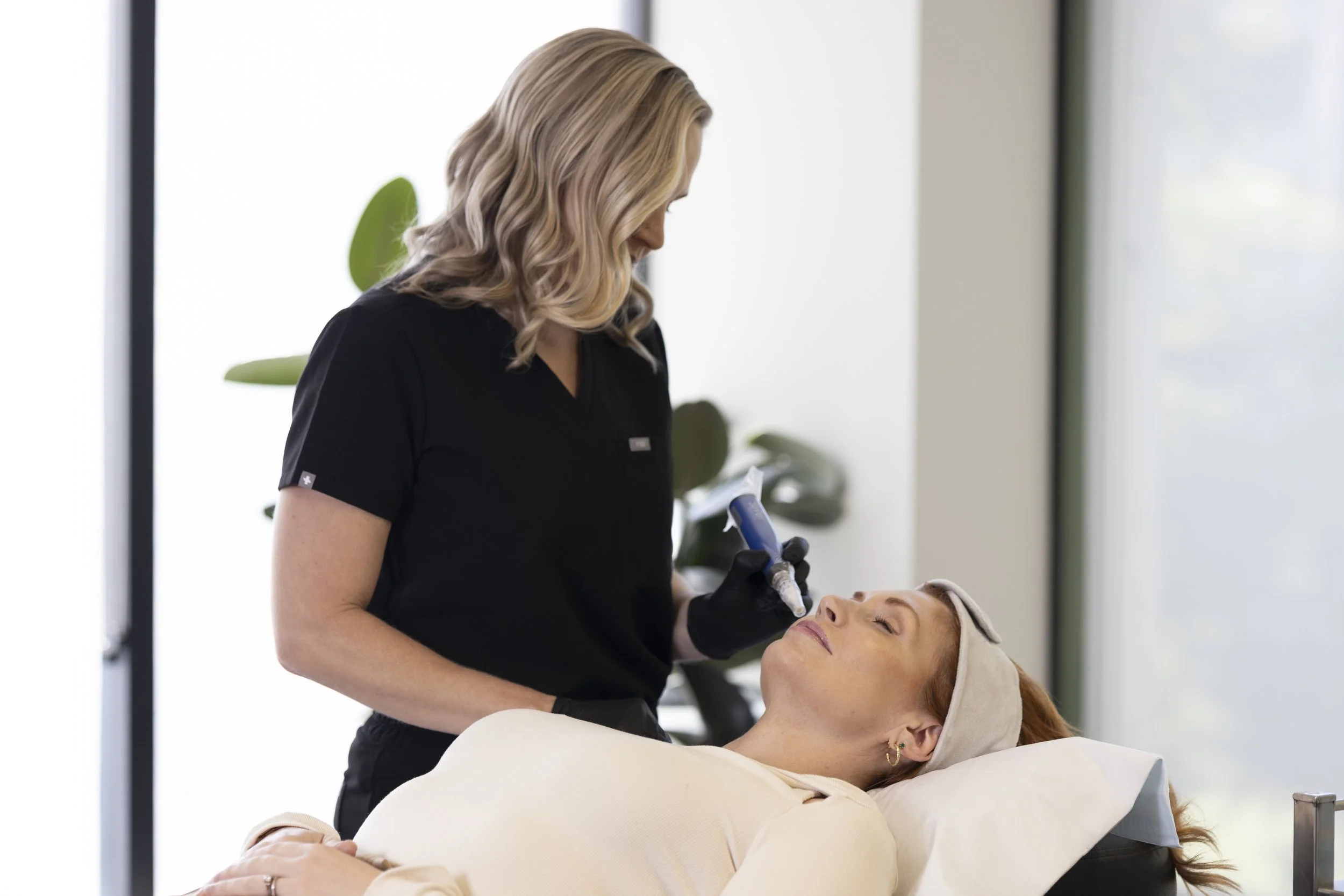 A woman receiving a cosmetic treatment from a professional esthetician in a clinic, lying on a treatment bed with her eyes closed.