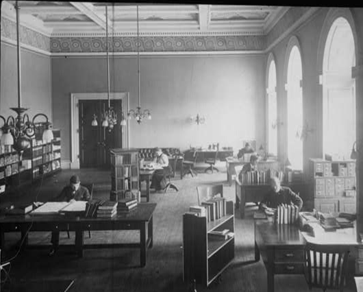 A black and white photo of a library reading room with several people reading and working at desks, large windows, bookshelves, and a high ceiling.
