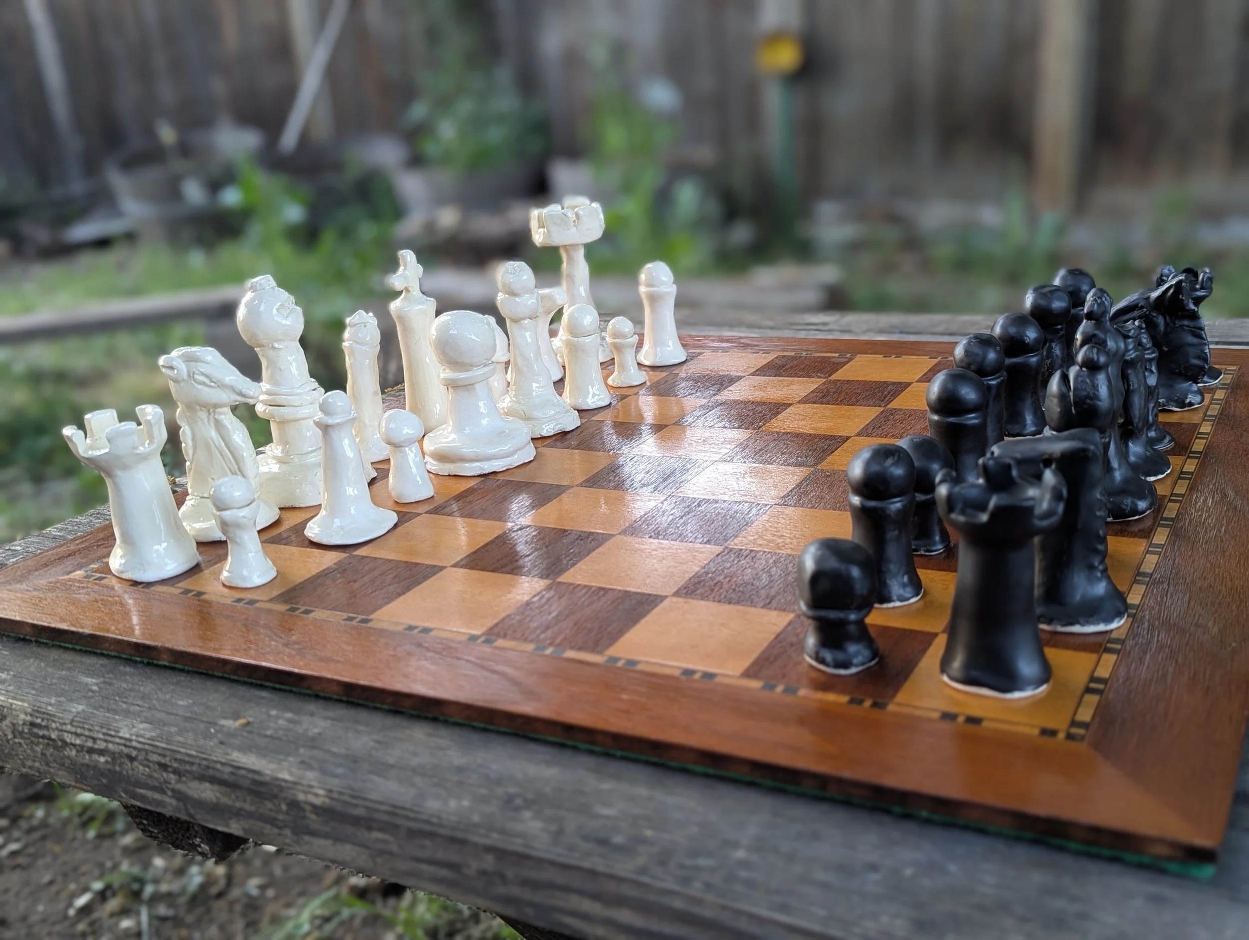 Black and white hand made ceramics chess pieces sit on wooden chess board. The pieces are not standard sizes.