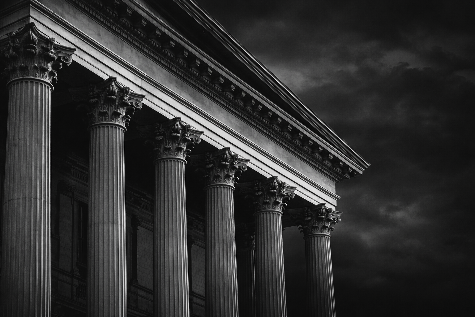 Black and white photo of classical building with tall Corinthian columns and ornate architectural details, dark cloudy sky in background.