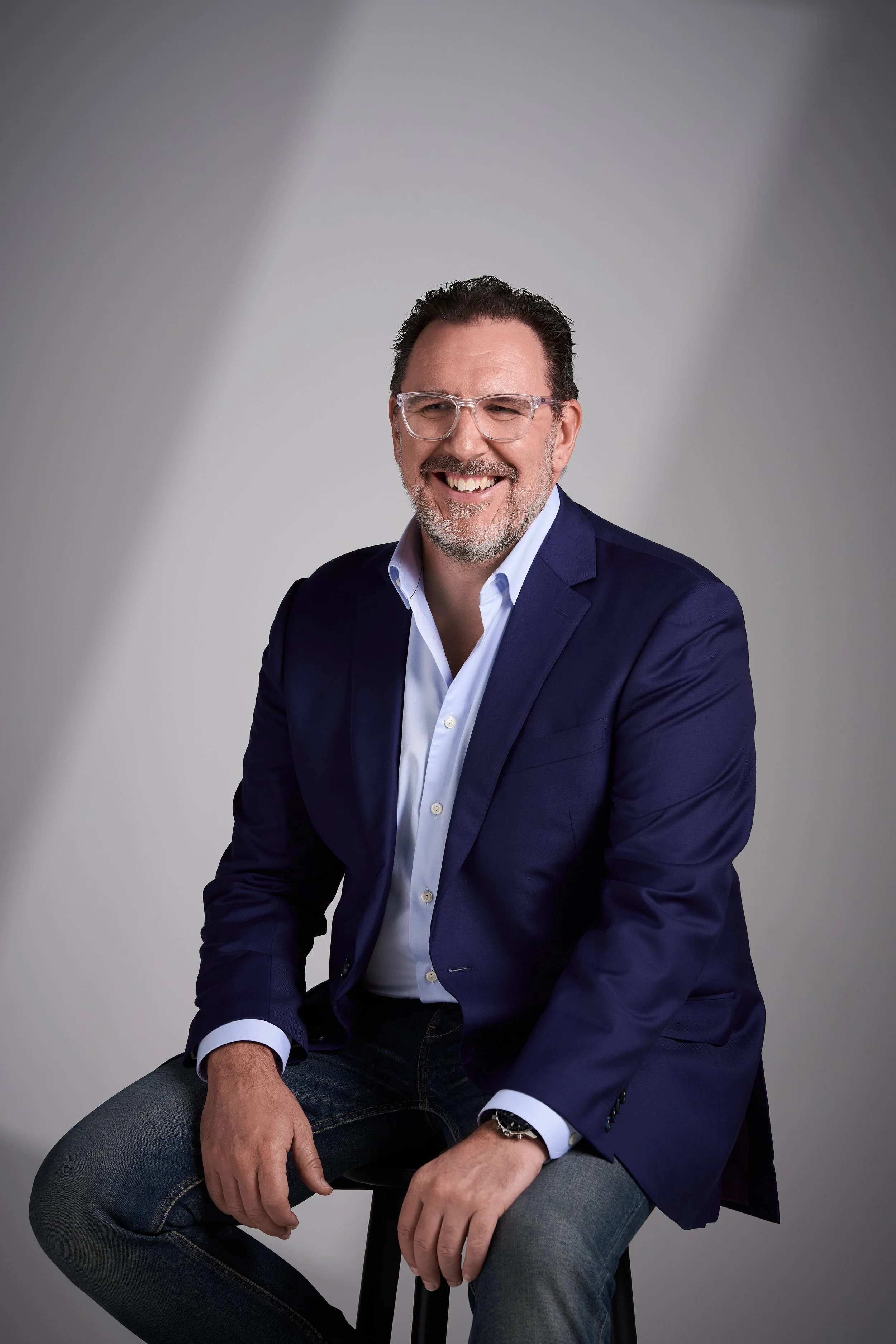 A smiling man with glasses, short dark hair, wearing a navy blazer, white shirt, and jeans, sitting on a stool against a plain light gray background.