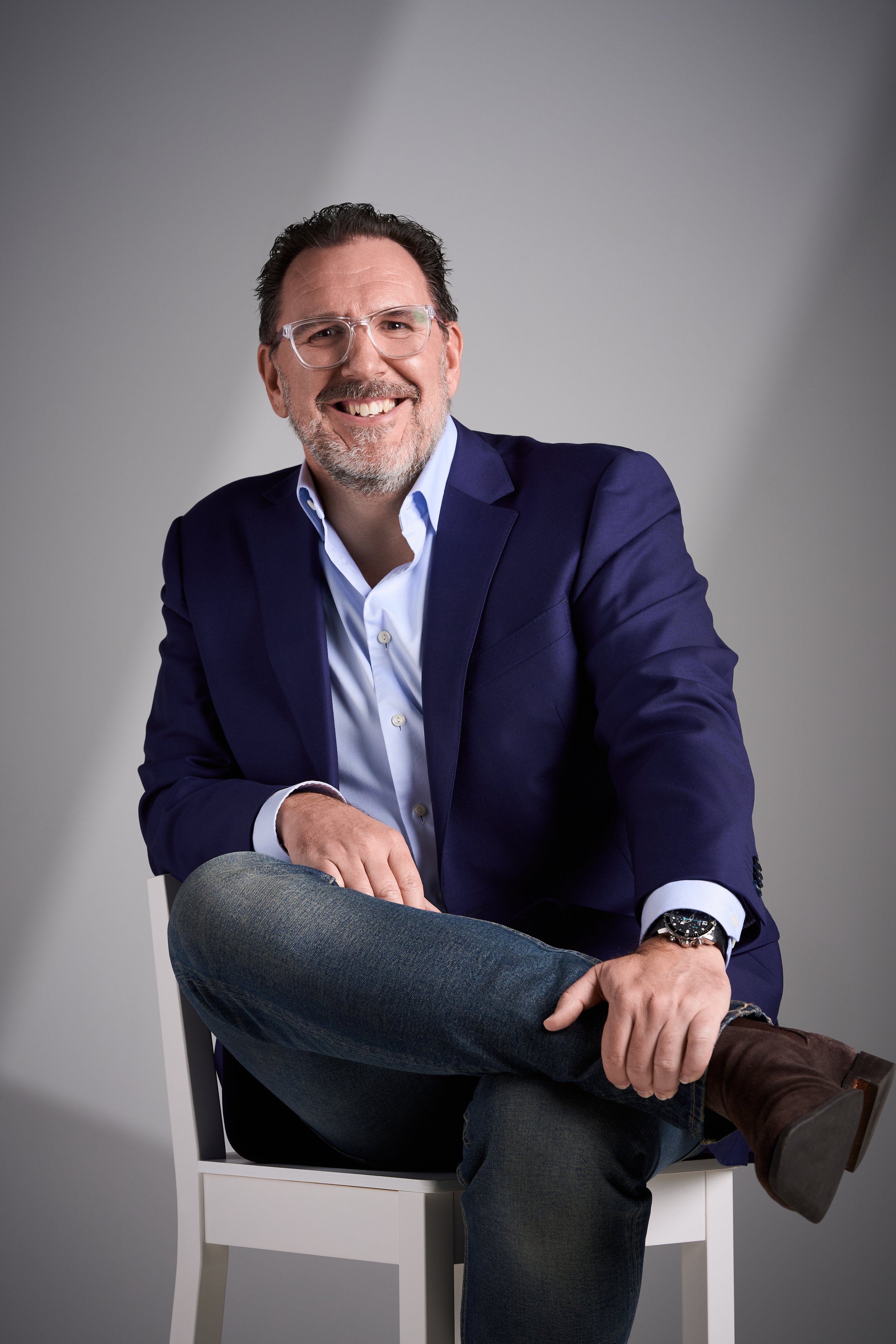Scott Greatrex with glasses and a beard smiling, wearing a navy blazer, light blue shirt, dark jeans, brown boots, and a watch, sitting on a white chair against a gray background.