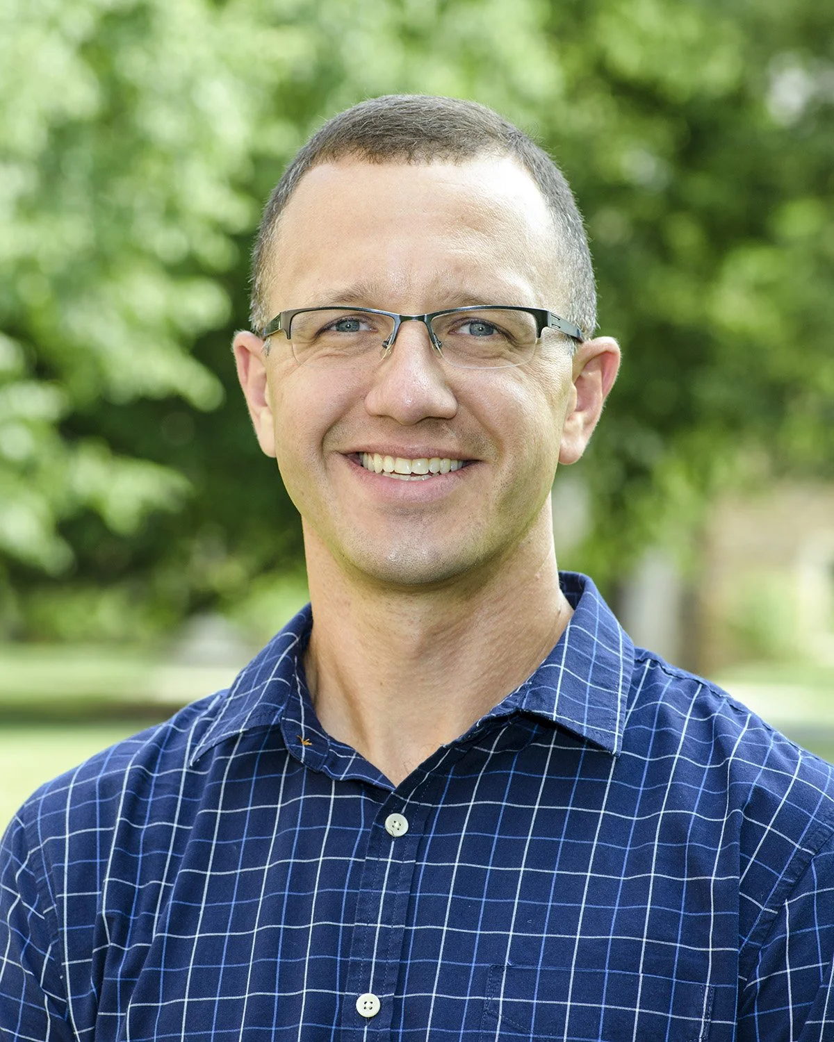 A smiling man with short brown hair, glasses, and a blue checkered shirt standing outdoors in front of a blurred green background.
