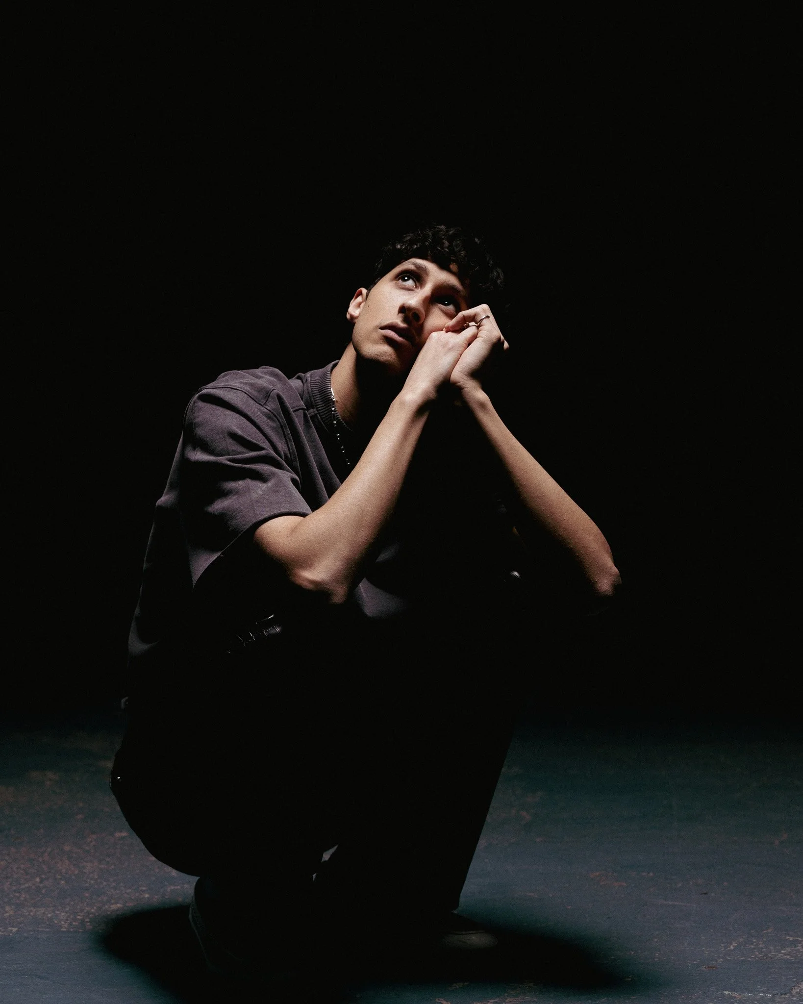A young man crouching in a dark space, looking upward with a thoughtful expression, hands clasped near his face.