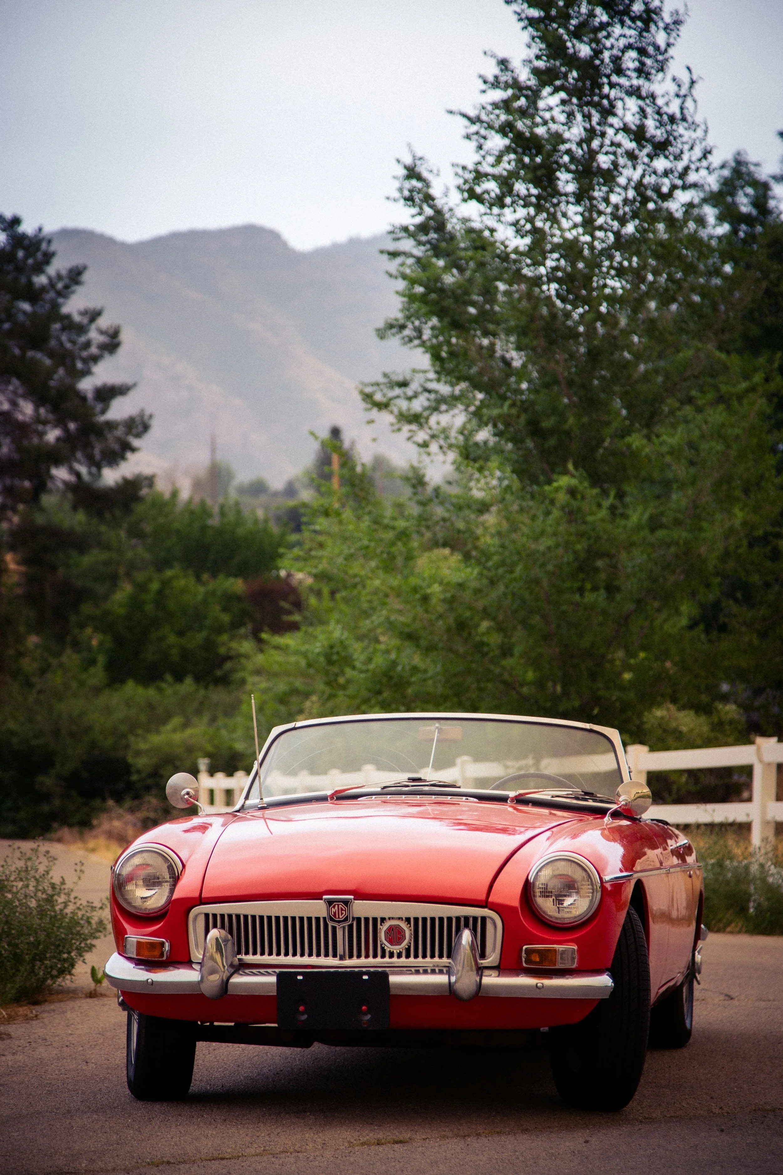 Front view of a vintage red MG convertible car parked on a paved driveway, with a white fence, green trees, and mountainous landscape in the background.