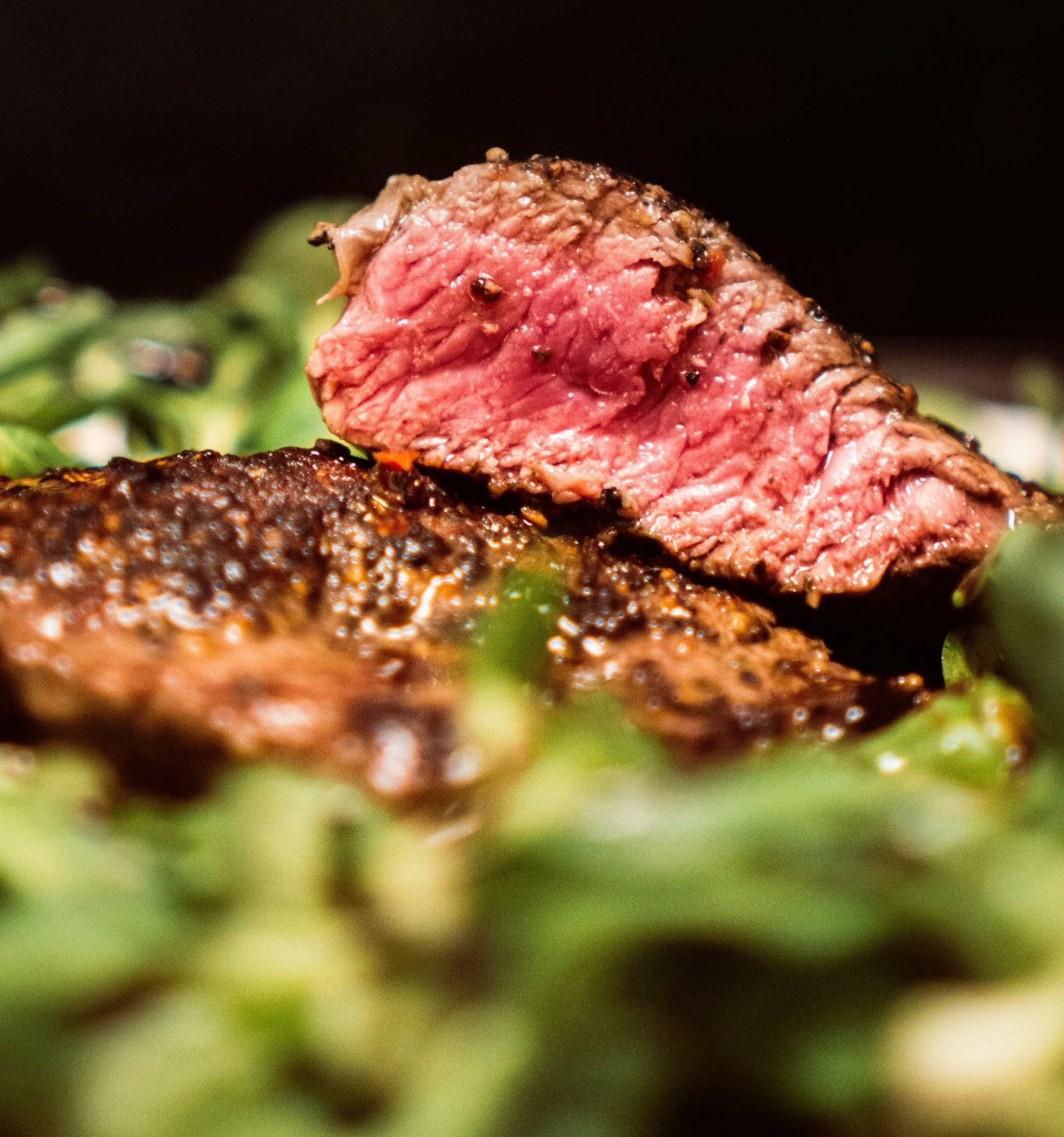 Close-up of a cooked, medium-rare steak with a crispy, seasoned exterior and pink center, resting on a bed of greens.