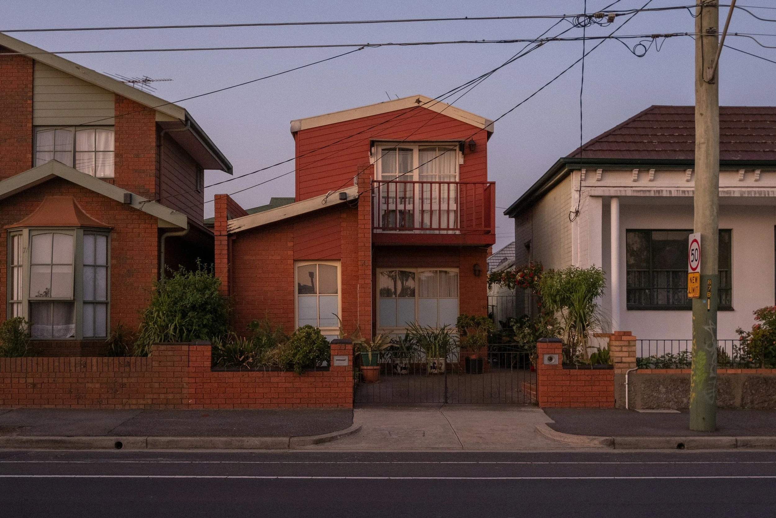 Three residential houses with brick and wood exteriors, a brick fence, a sidewalk, power lines above, and a street in the foreground at dusk.