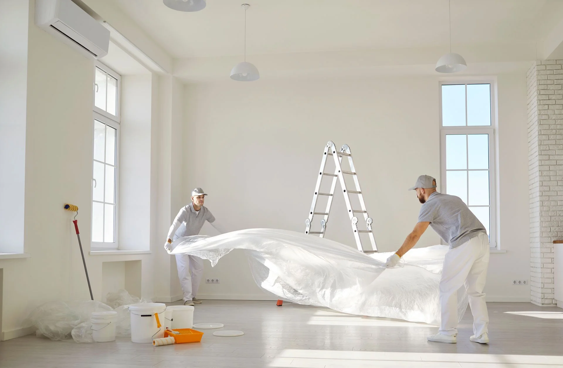 Two workers in white clothes are laying down a large white sheet in a bright, modern room with large windows and white walls. There is a ladder, buckets, paint rollers, and other decorating tools around them, indicating they are preparing for painting or renovation.