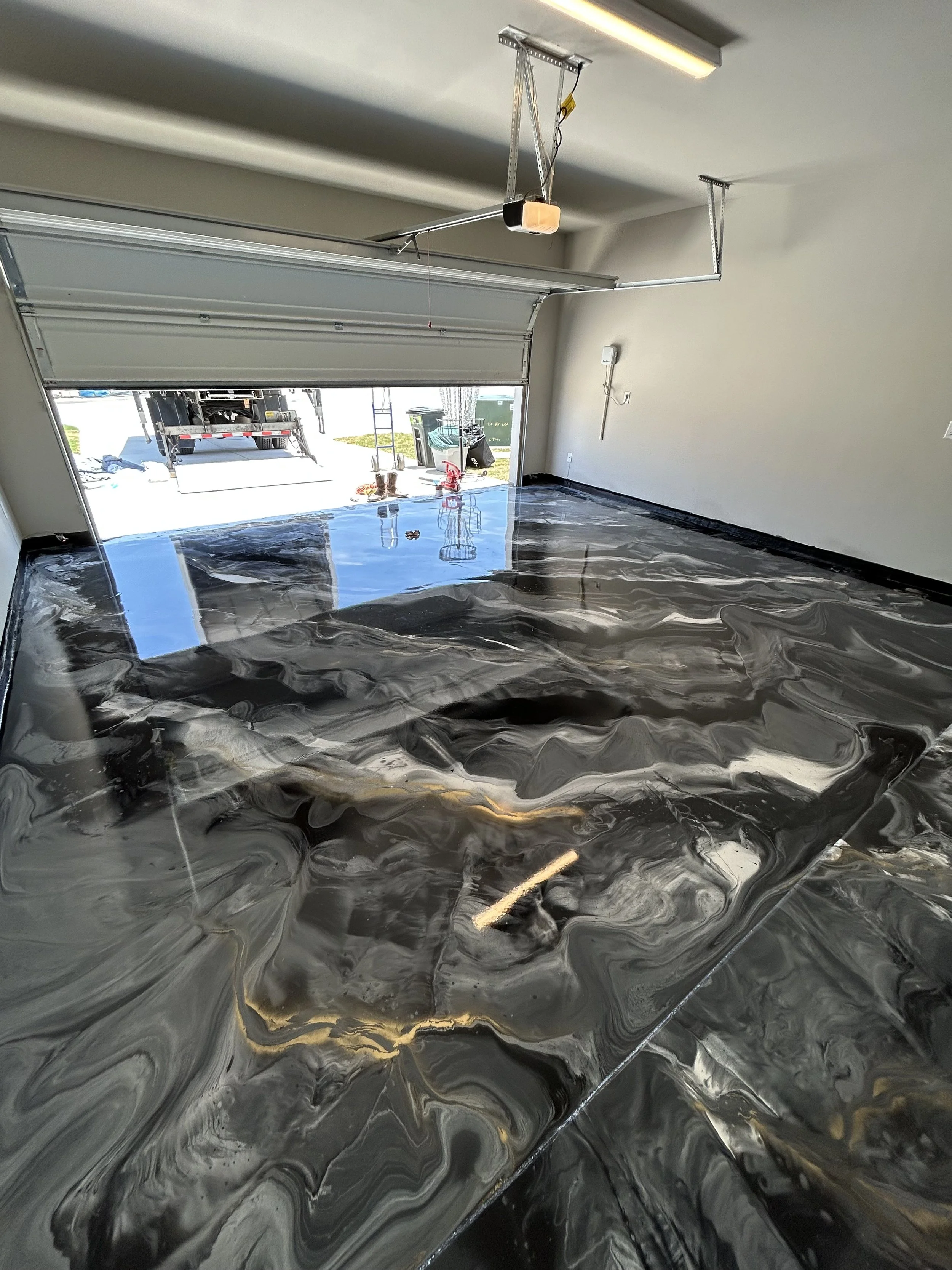 Empty garage with a shiny, black marble-like floor and an open garage door leading outside. There are some tools and a trash bin outside.