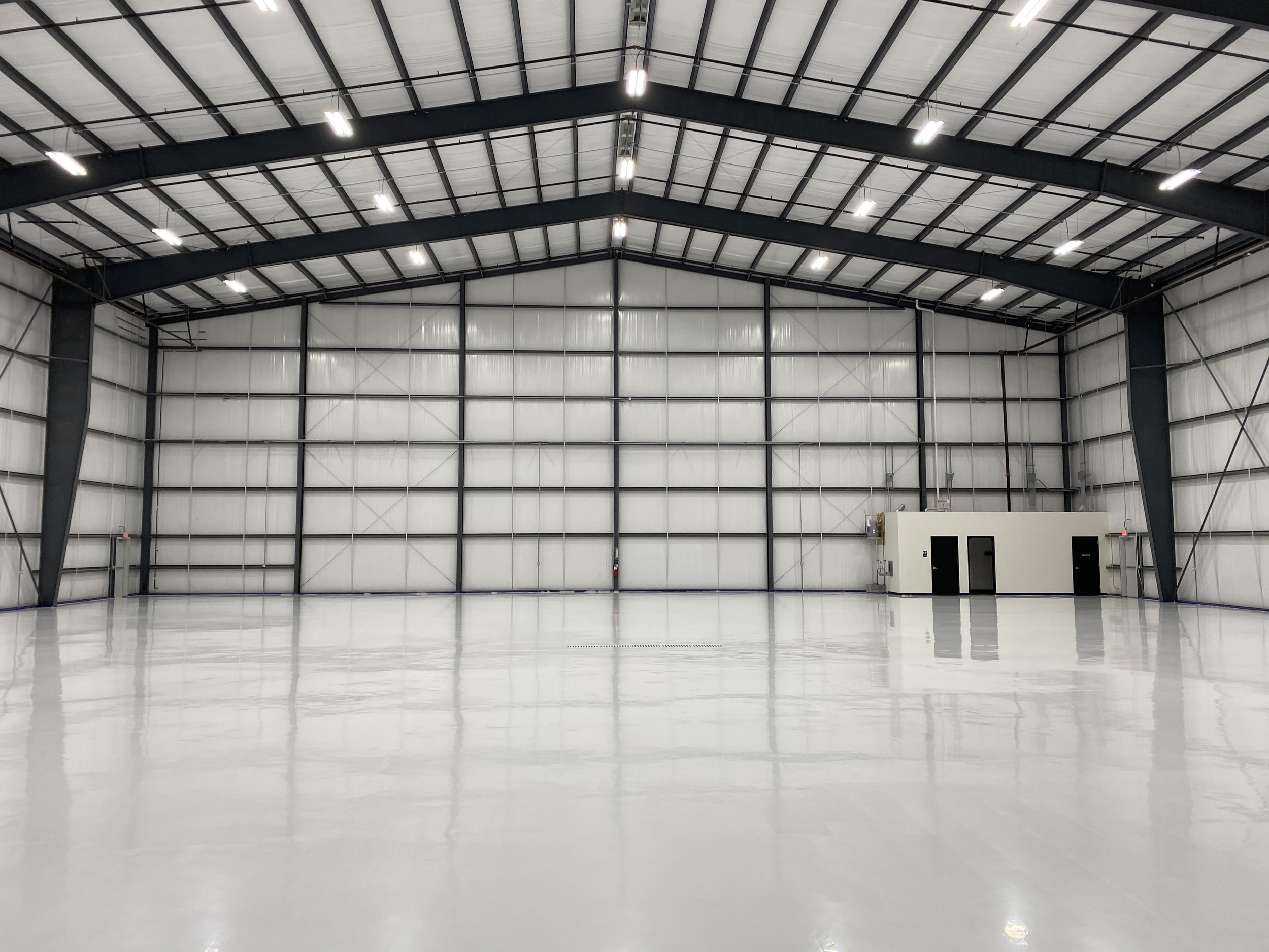 Empty industrial warehouse with polished concrete floor, high ceiling with metal beams, and bright overhead lighting.