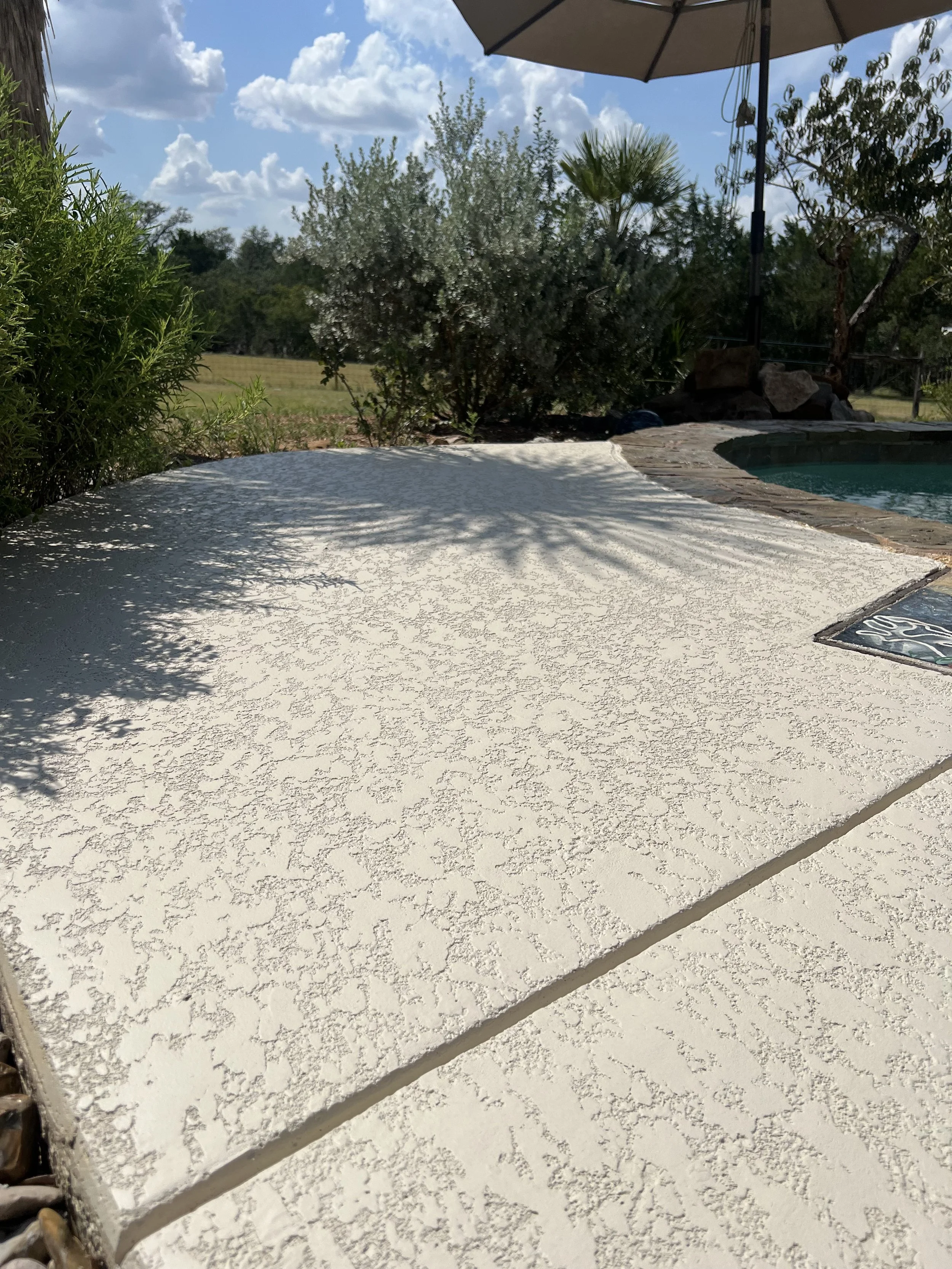 Close-up of a textured, off-white concrete pool deck with shadows of nearby plants. In the background, part of a swimming pool with a stone border, outdoor umbrella, and various trees and bushes under a partly cloudy sky.