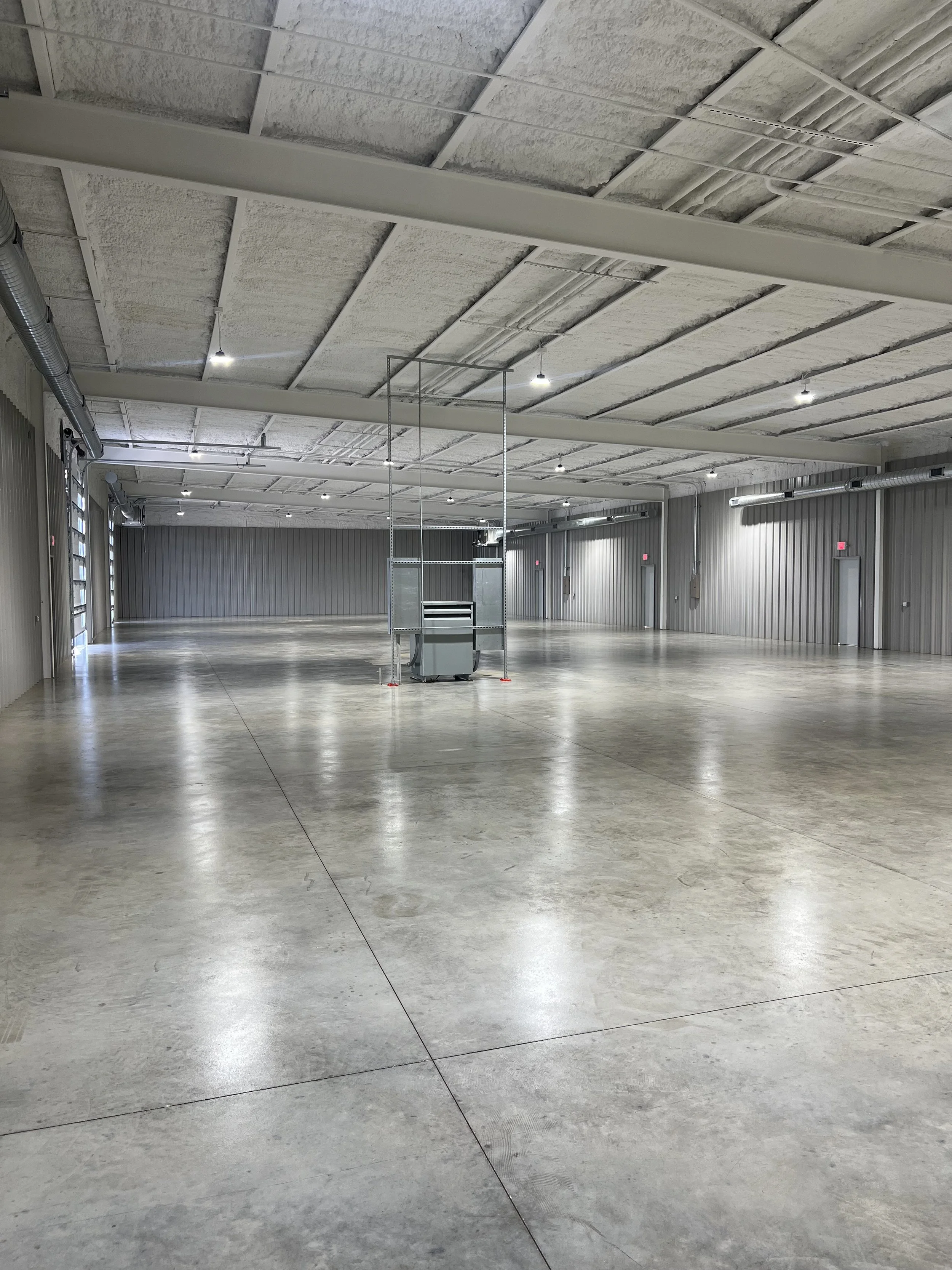 Empty industrial warehouse with concrete floors, metal walls, and exposed ceiling with lighting and ventilation ducts.