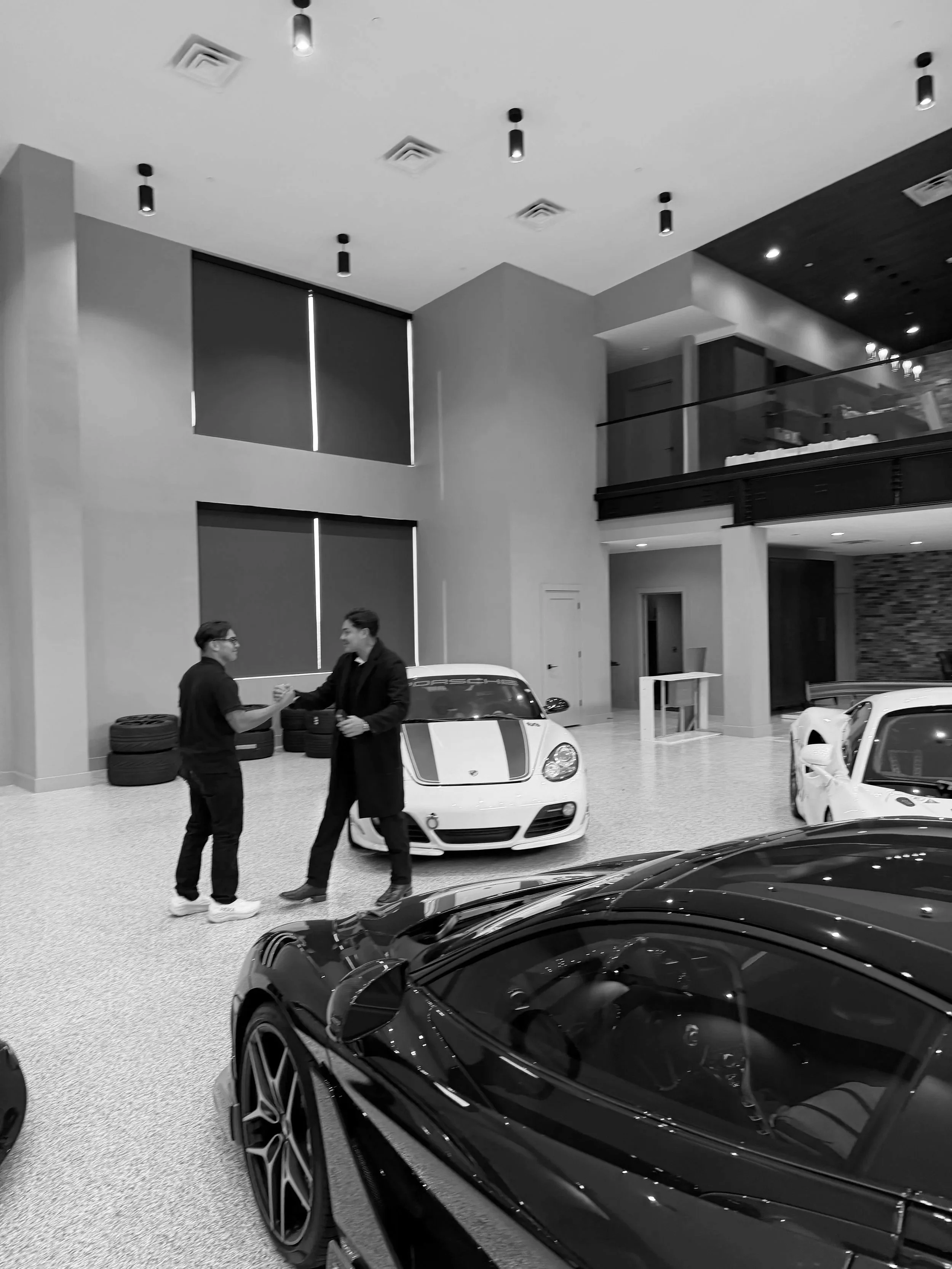 Two men shaking hands next to a white sports car inside a car dealership or showroom, with other luxury cars visible in the foreground and background.