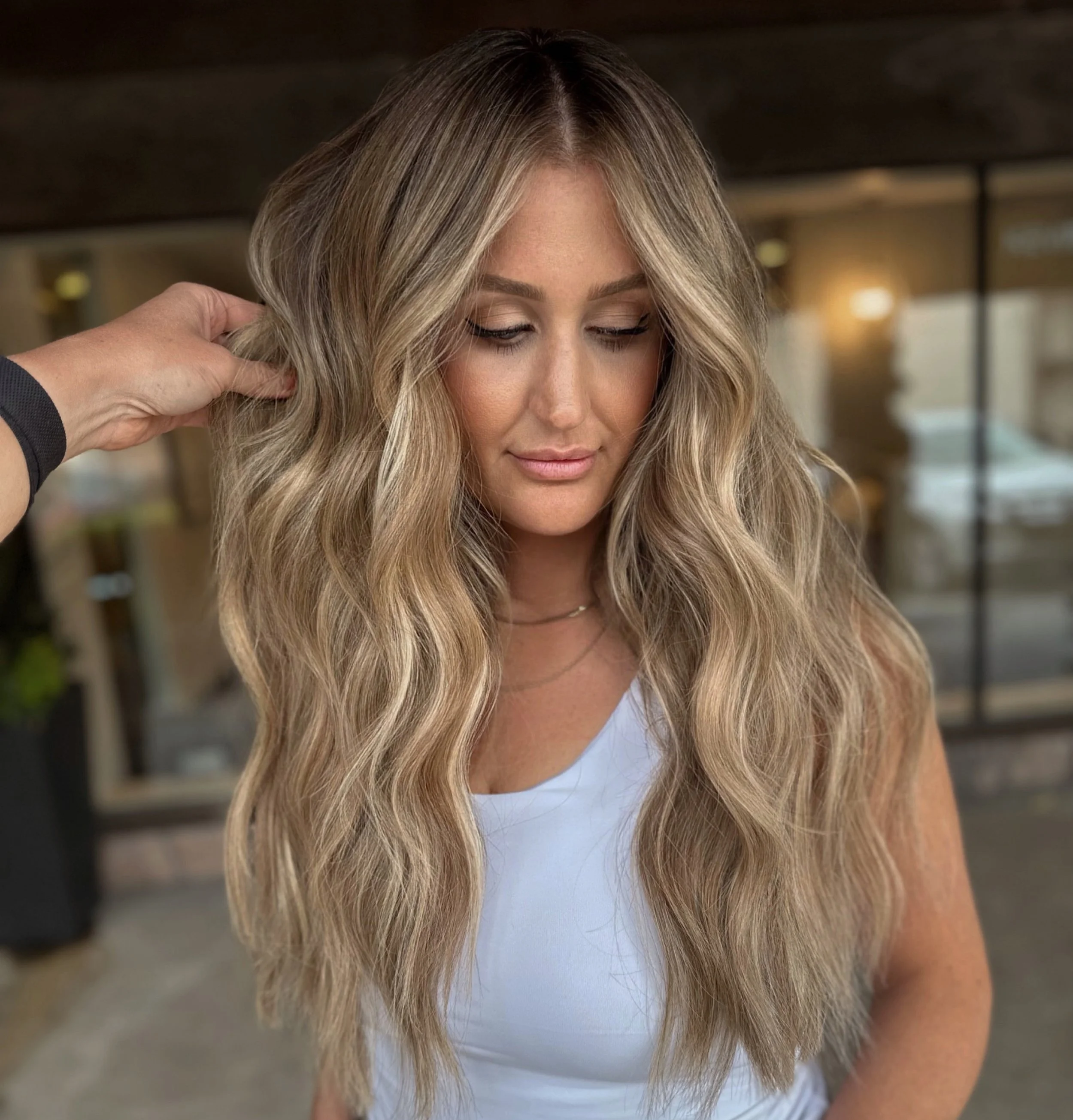 A woman with long, wavy blonde hair and a light makeup look, wearing a white top, being styled by a person's hand on her hair in an indoor setting.