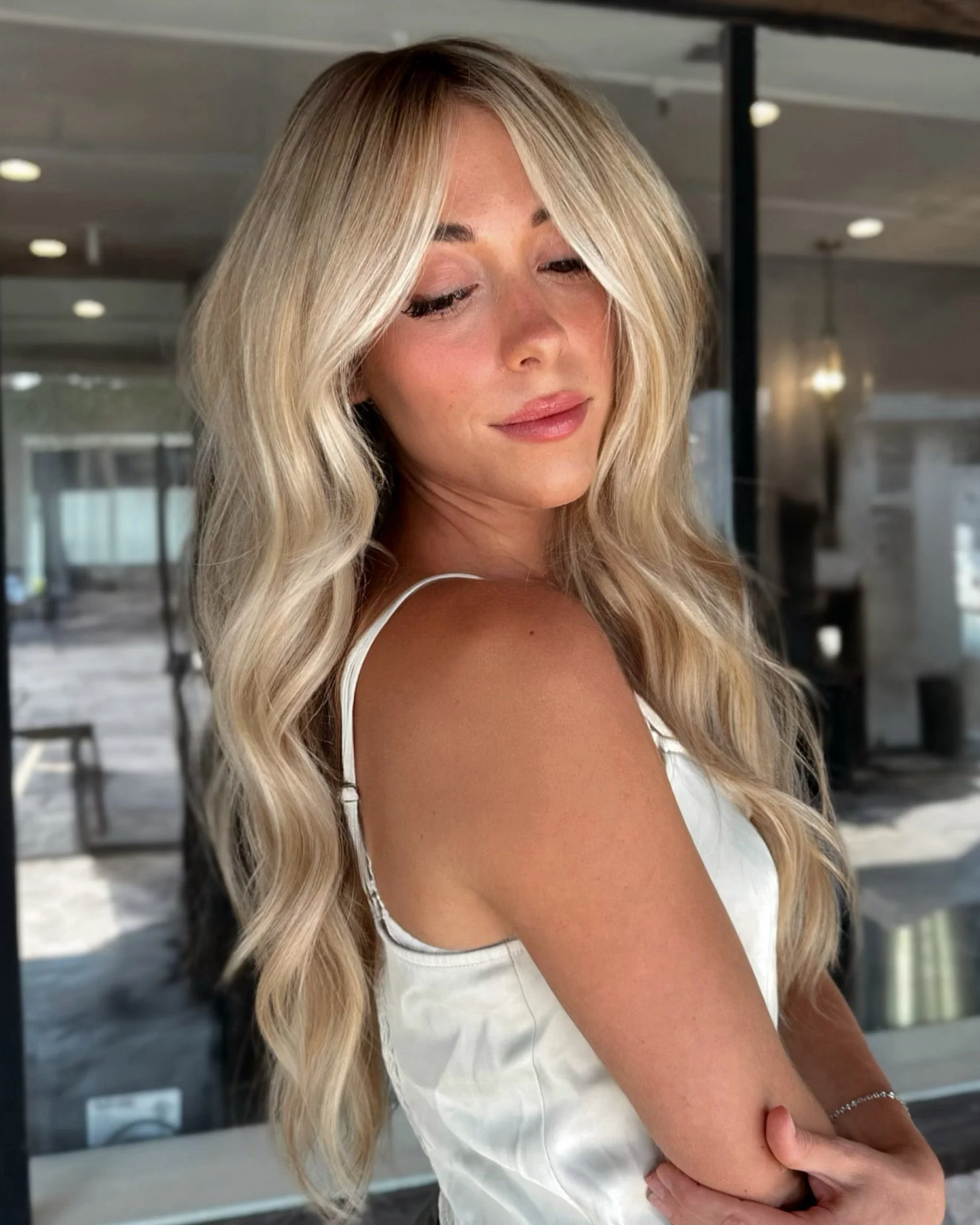 A woman with long wavy blonde hair wearing a white sleeveless top, standing indoors near a window with black frames and ceiling lights, looking down with a slight smile.
