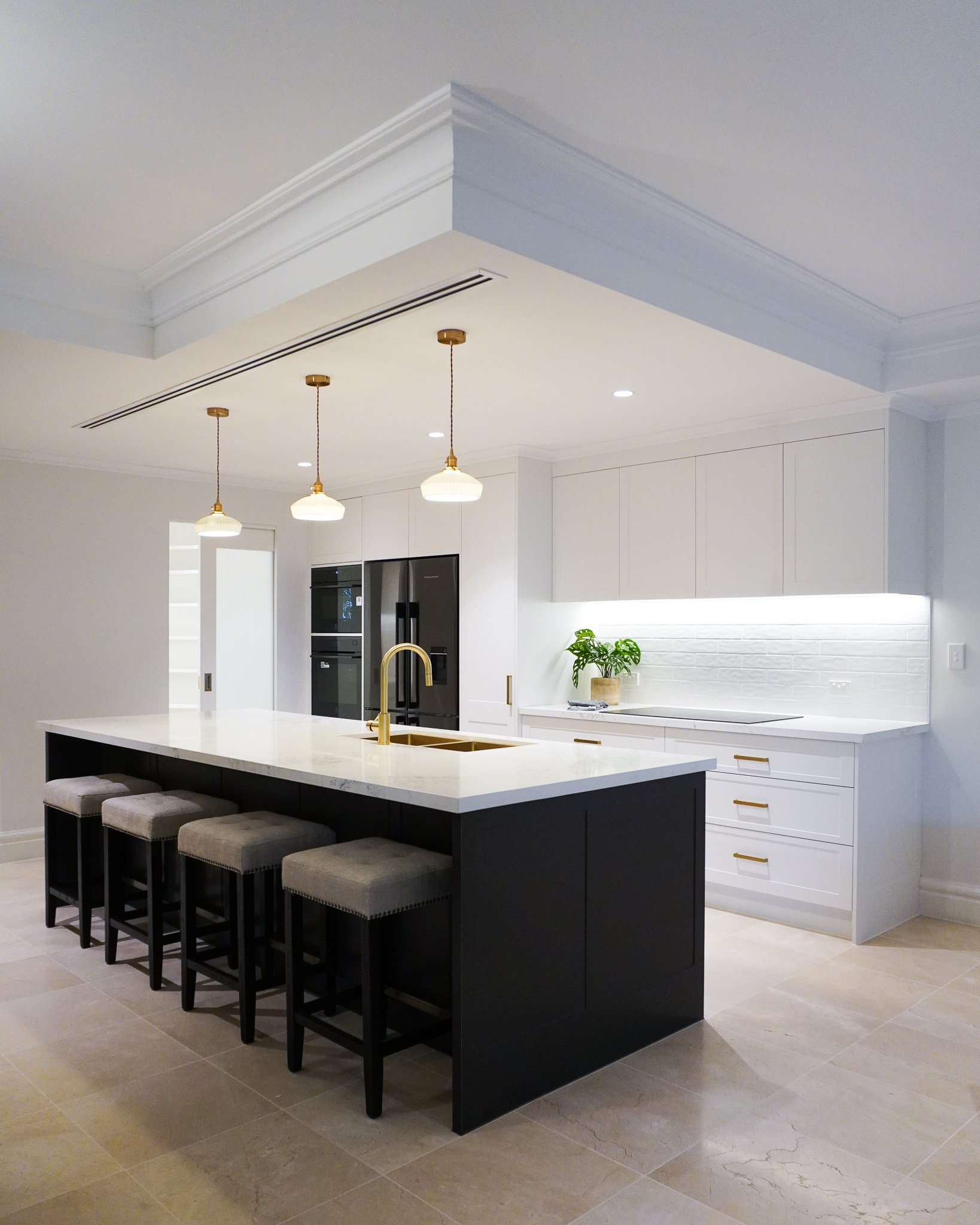 Modern kitchen with white cabinets, black refrigerator, black island with gold faucet, beige stools, and hanging pendant lights.