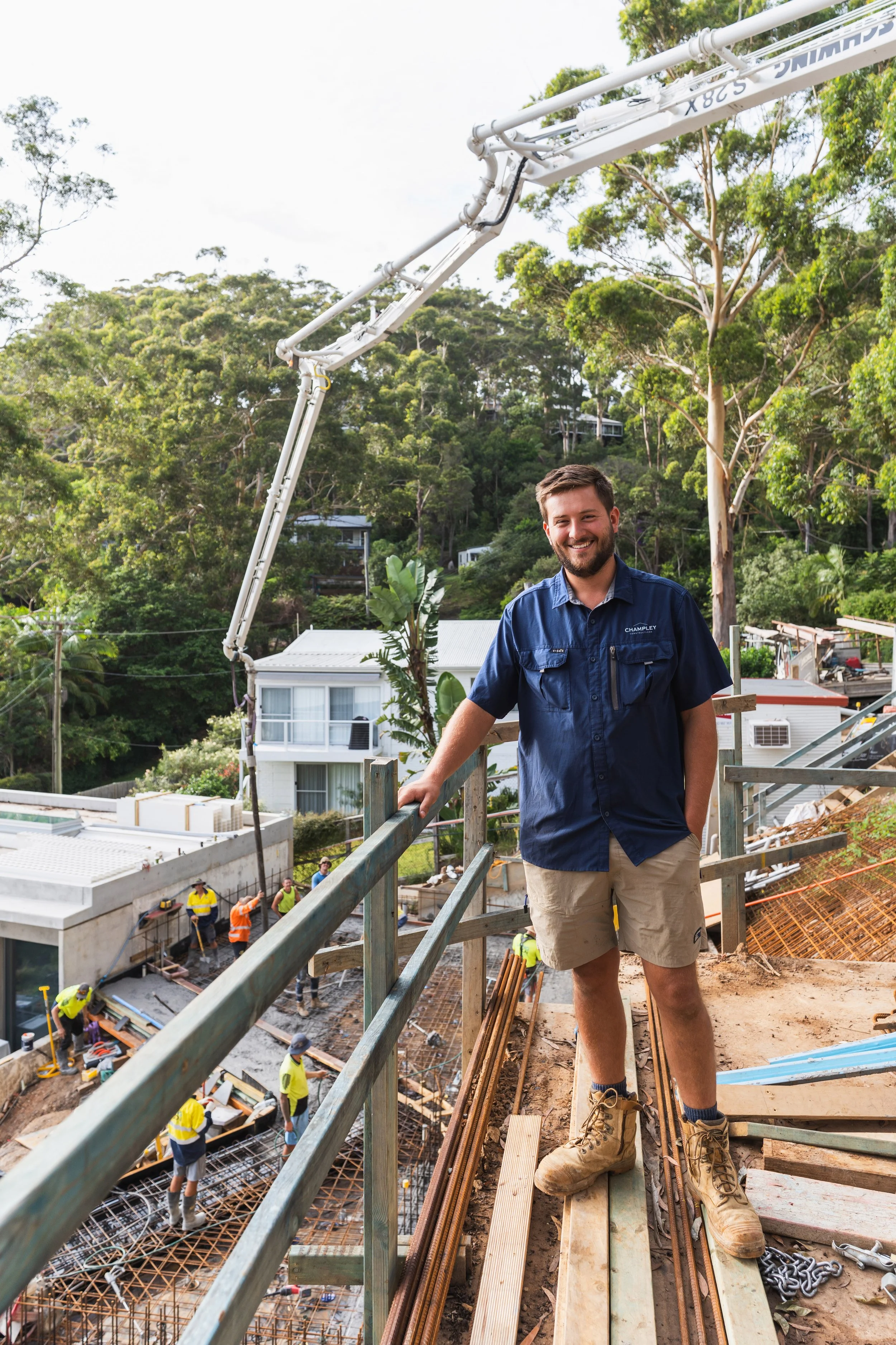 A construction worker smiling while standing on a construction site overlooking workers below and a green hillside in the background.
