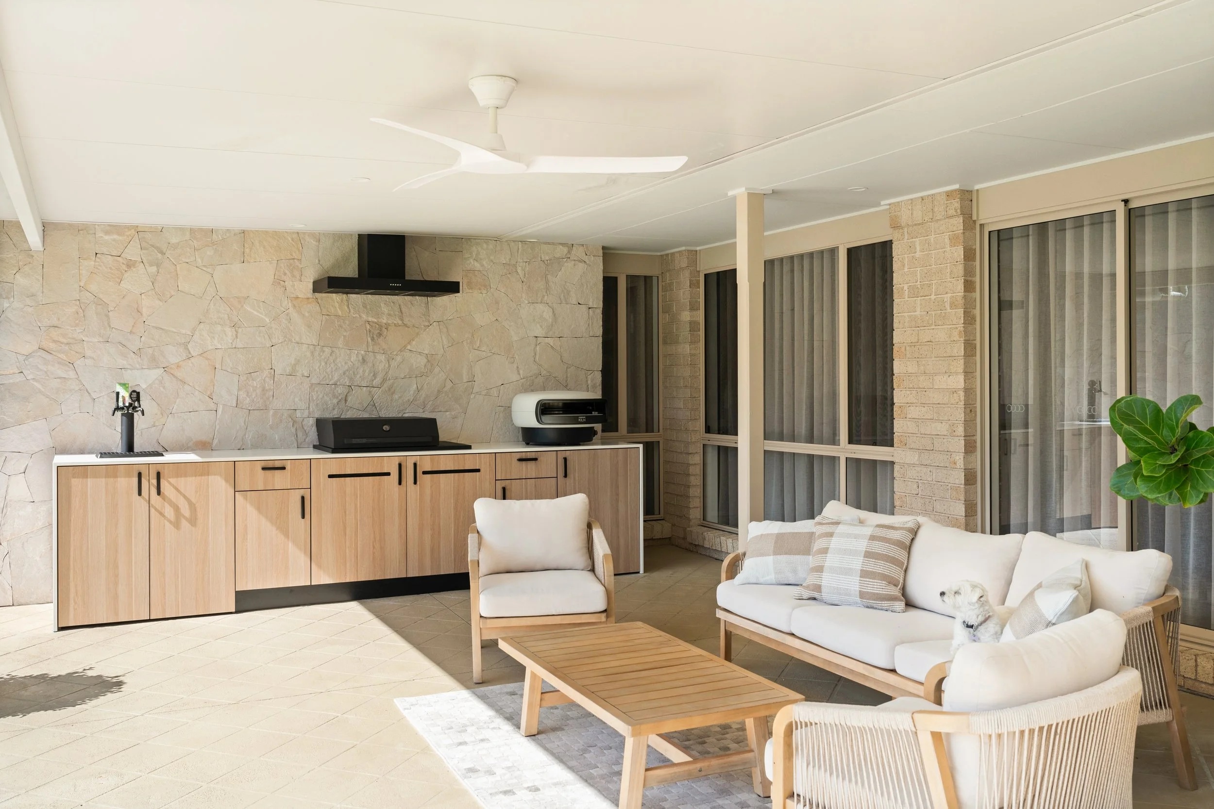 Sunlit outdoor patio with beige tile flooring, a white sofa with striped and plain cushions, matching armchair, wooden coffee table, built-in wooden kitchen cabinets, modern countertop appliances, large windows with vertical blinds, a ceiling fan, and a potted plant.