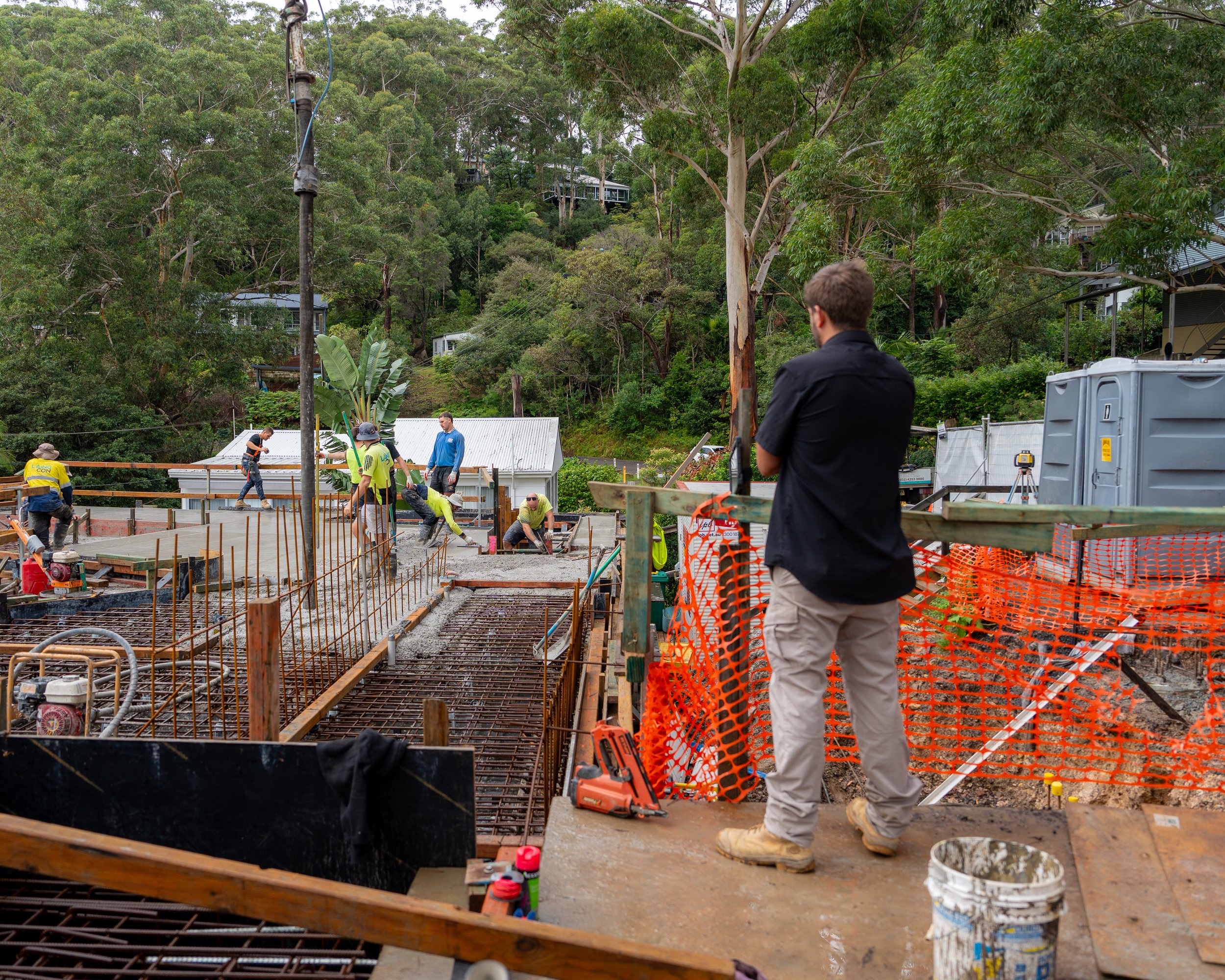 Construction workers on a job site building a structure with steel rebar framework, surrounded by orange safety fencing, in a wooded area with trees and buildings in the background. A man in a black shirt and khaki pants stands observing the work.