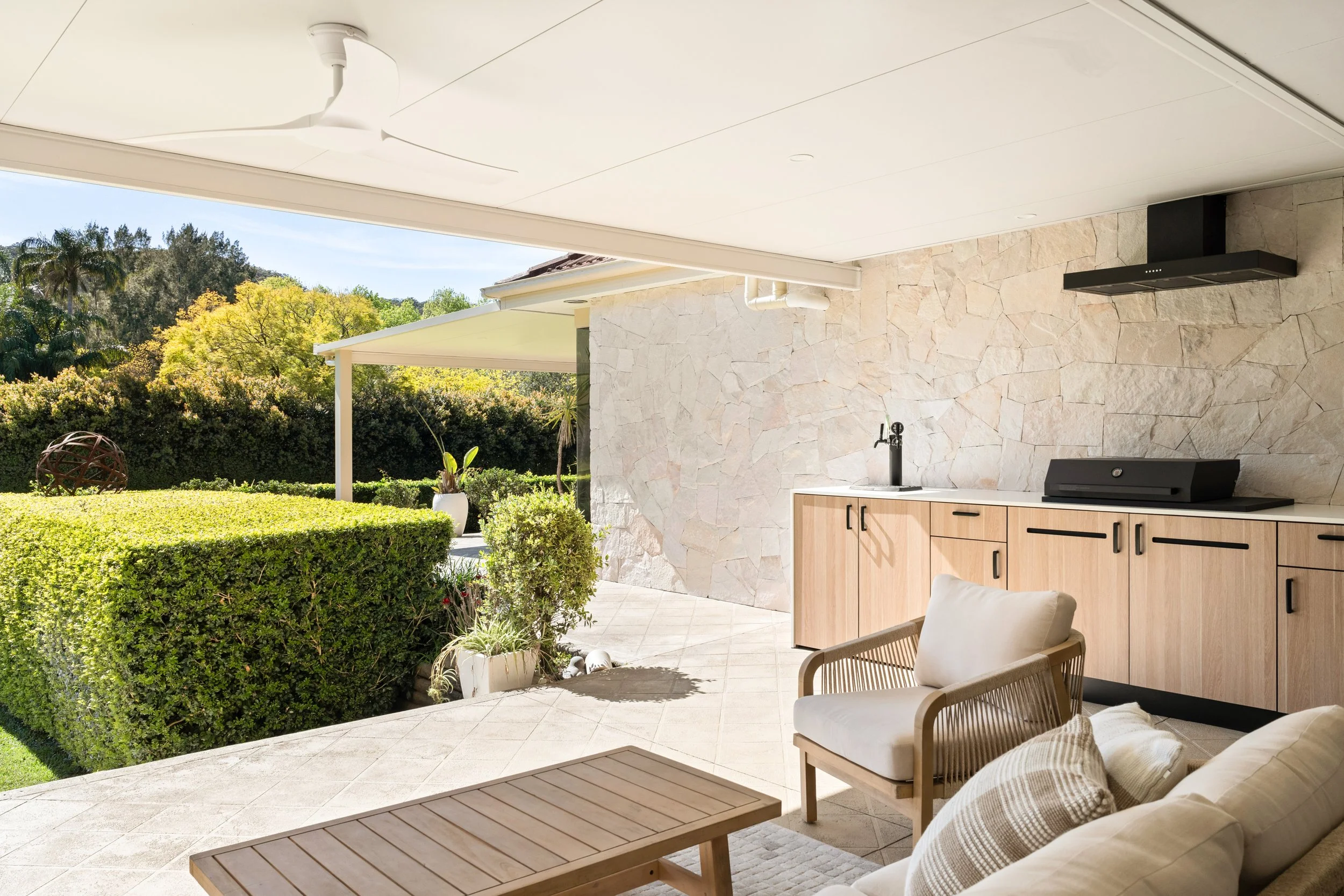 Outdoor patio with beige tile flooring, wooden cabinetry with black handles, a black grill, a black faucet, lush green bushes, a potted plant, a lounge chair with a wood frame and white cushion, and a sofa with beige cushions, overlooking a garden with trees.