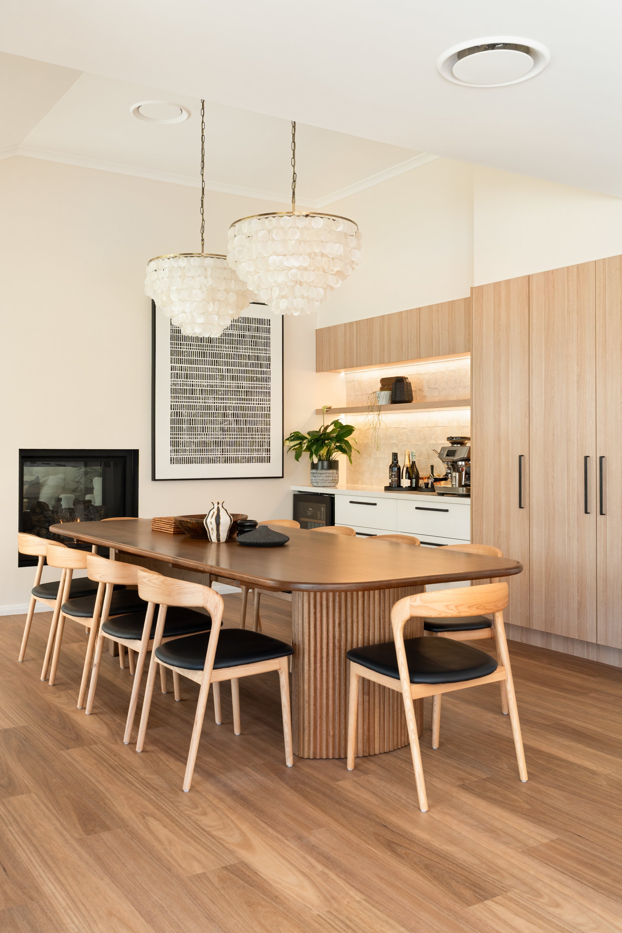 Modern dining room with a wooden table and eight black-cushioned chairs, a built-in cabinetry with shelf lighting, a large artwork on the wall, and two hanging chandeliers.