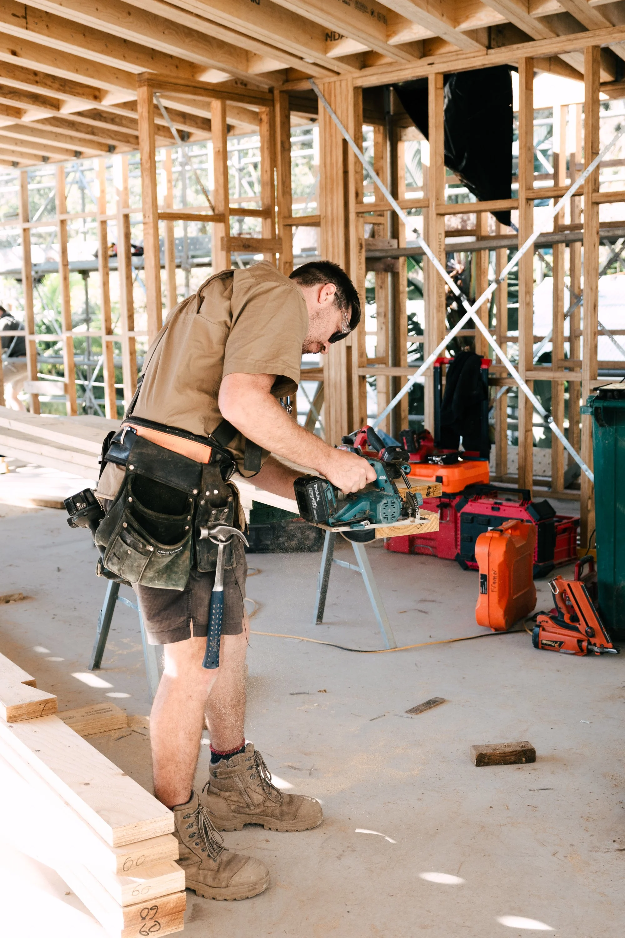 A man wearing work boots, shorts, and a tool belt is using a saw on a piece of wood at a construction site with a wooden framework. There are power tools and construction supplies around him.