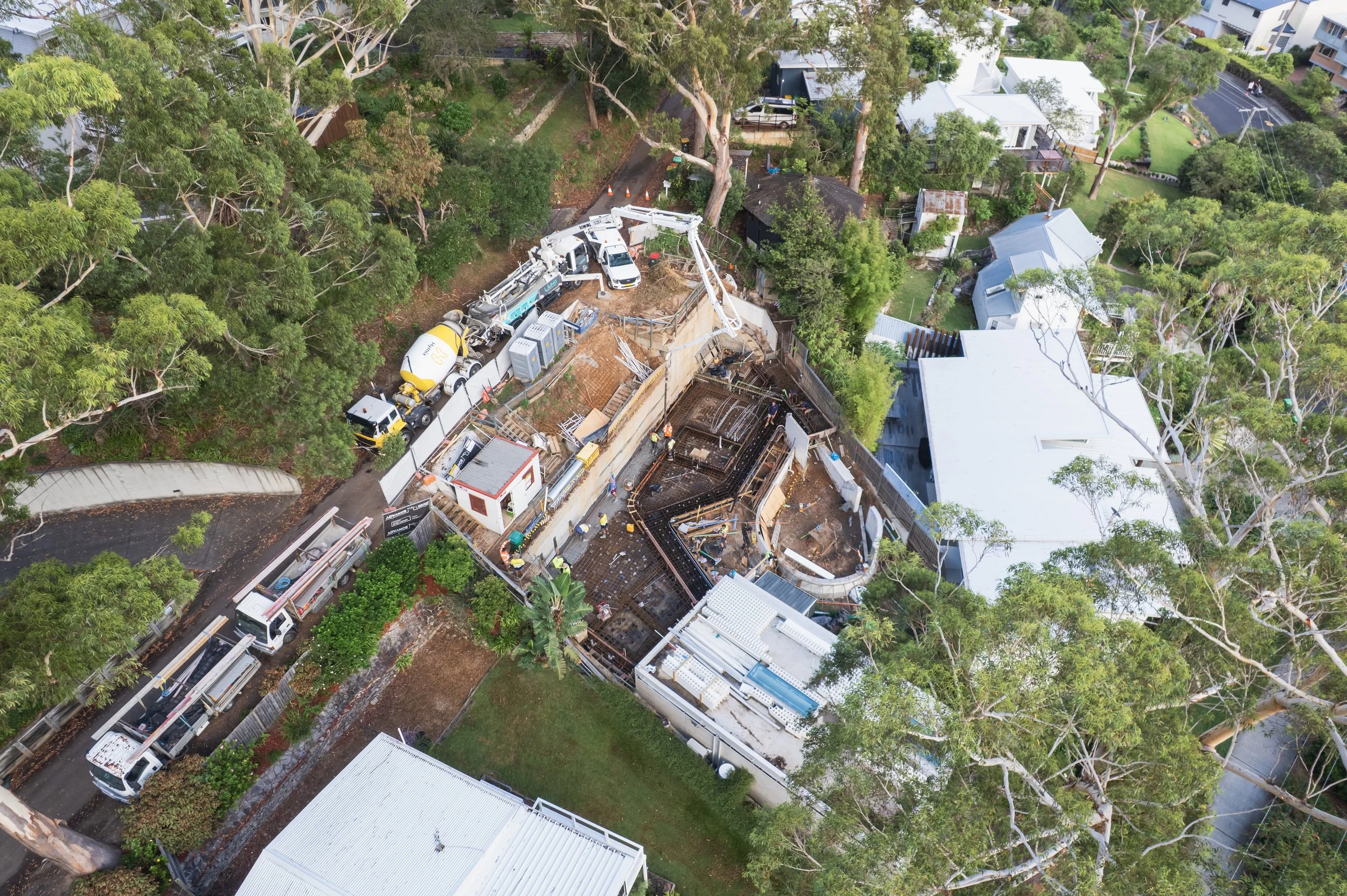 Aerial view of a construction site surrounded by trees and residential buildings, with construction vehicles and workers working on the foundation of a building.
