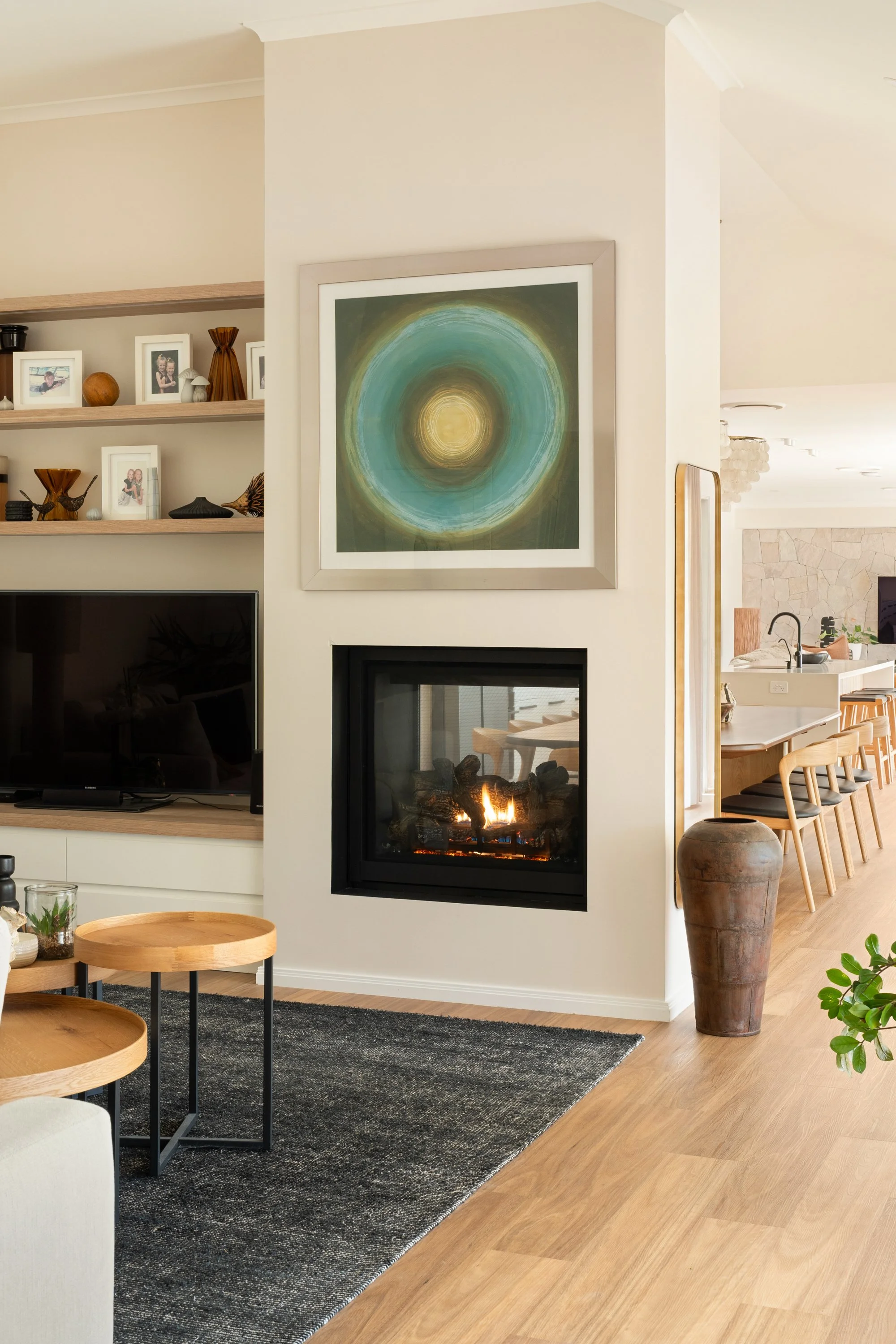 Living room with a fireplace, abstract artwork above, TV on a shelf to the left, decorative shelves with framed photos and ornaments, and a wooden dining area in the background.