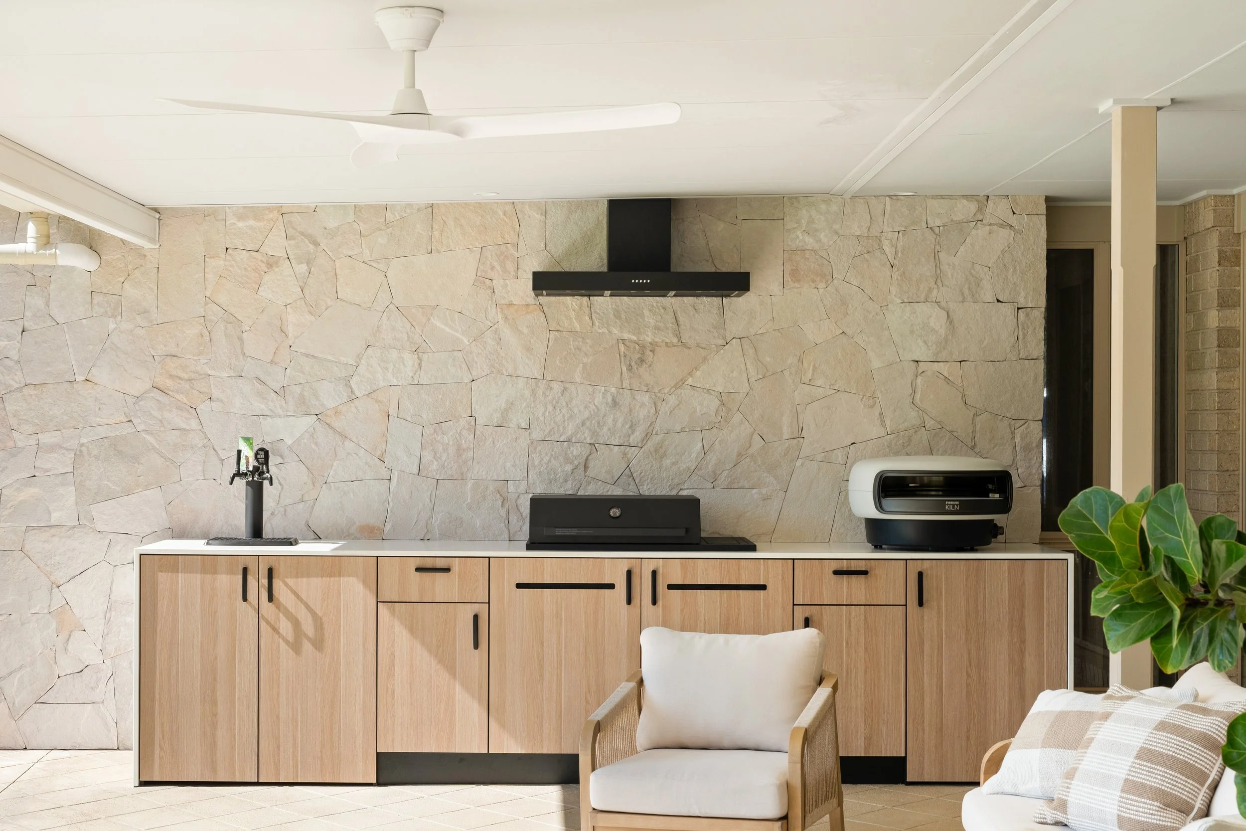 A modern kitchen with a stone accent wall, light wood cabinetry, and a white countertop. There is a black range hood, a white countertop appliance, a small black sink faucet, and a white chair with a pillow. A green plant is partially visible in the foreground.