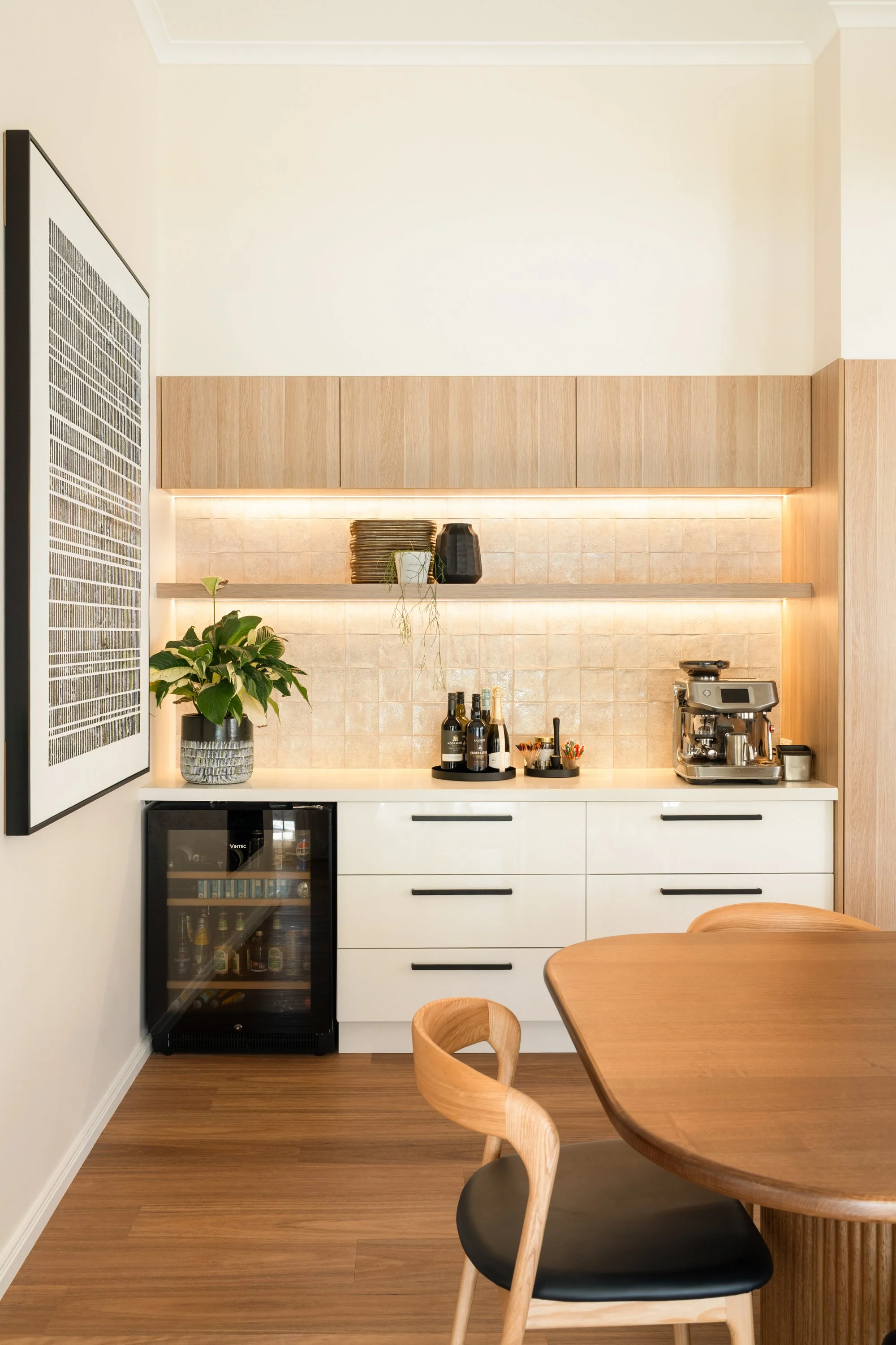 Modern kitchen with white drawers, a small wine fridge, a wooden table with chairs, a coffee machine, and decorative items on a backsplash