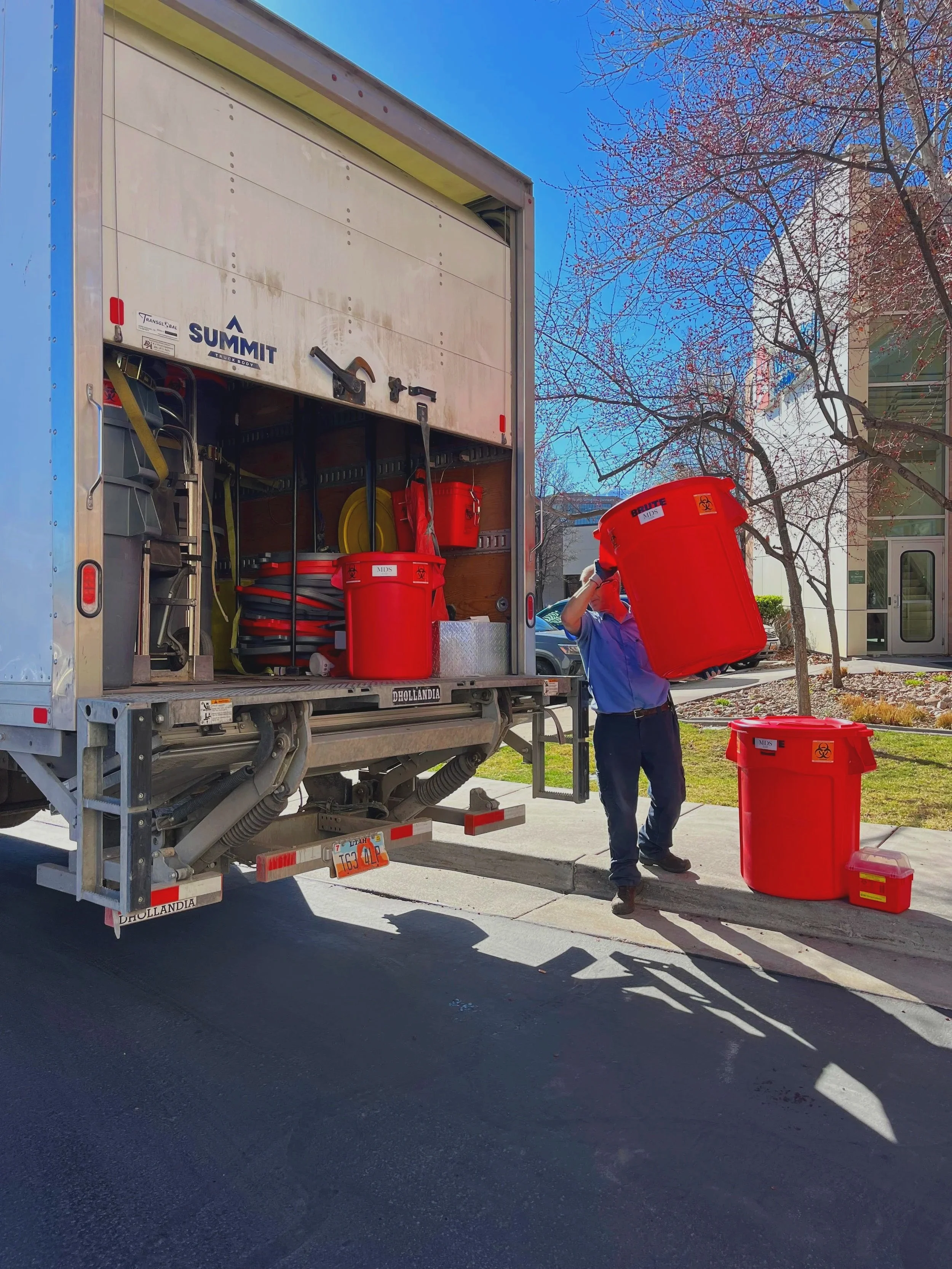 A person in a blue shirt and black pants loading a large red trash bin into a garbage truck on a sunny day, with trees and a building in the background.