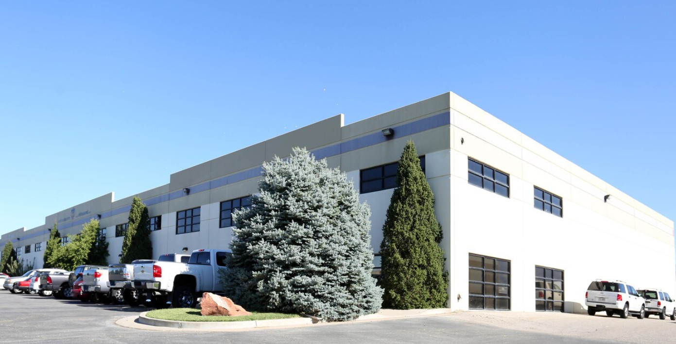 Large commercial building with white exterior, multiple dark windows, several parked cars and trucks, and landscaped bushes and trees in front, under a clear blue sky.