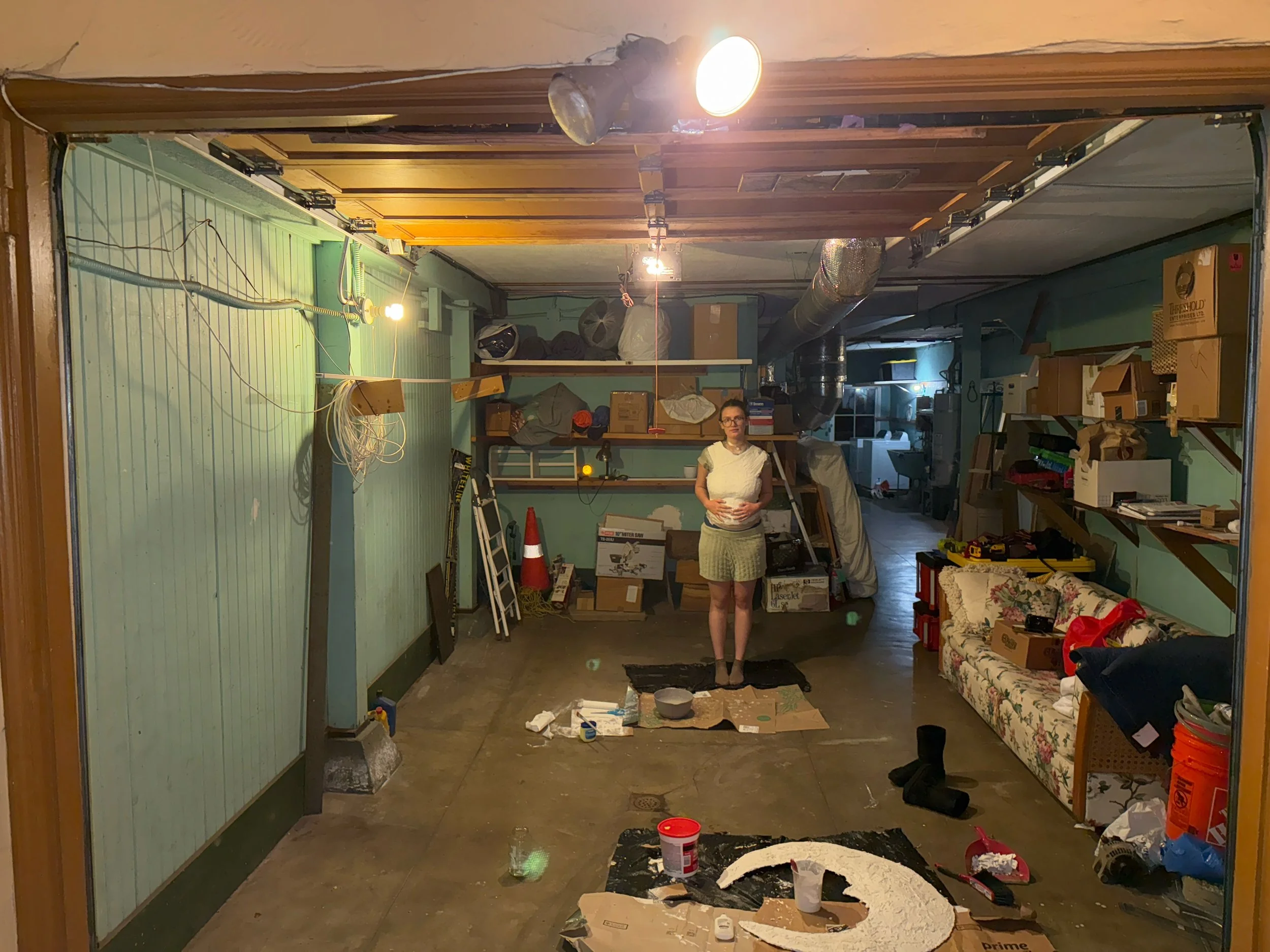A woman stands in the middle of a cluttered garage or workshop with various boxes, tools, and supplies. The space has painted walls, shelves filled with items, and a couch on the right side. Items like a traffic cone, ladder, and large ductwork are visible around the room, which appears to be in the process of organization or renovation.