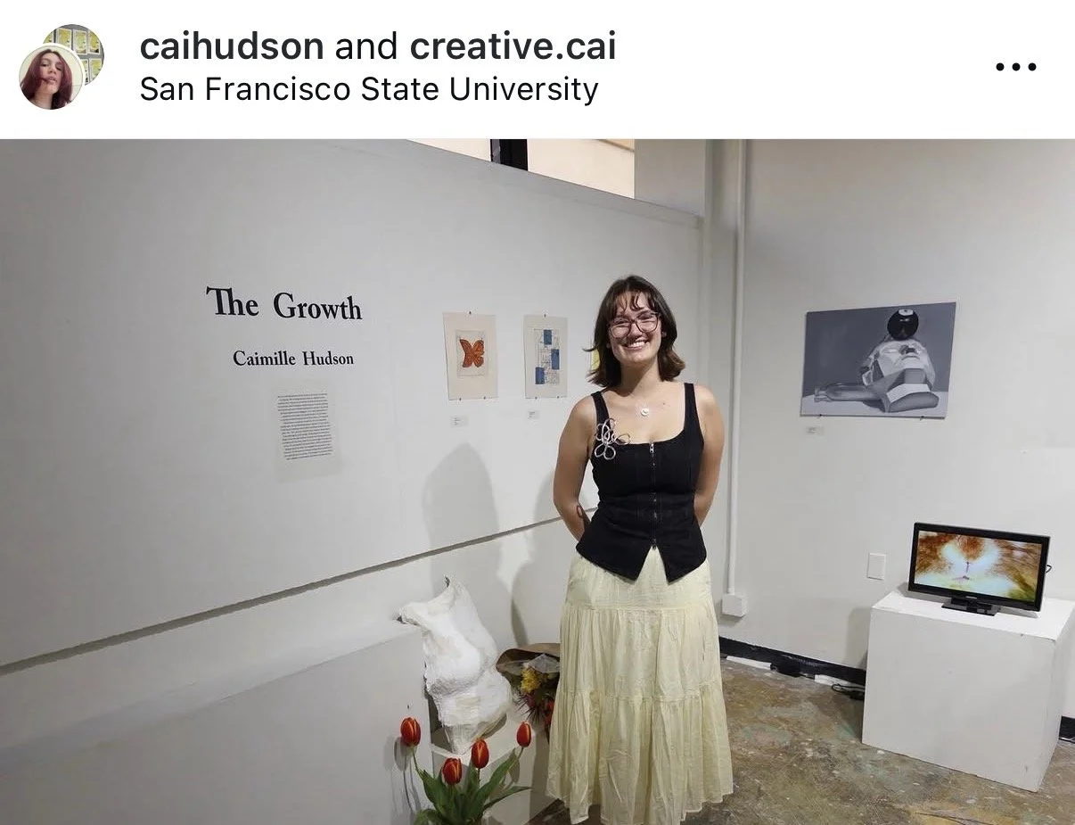A woman standing in an art gallery smiling, with a banner titled 'The Growth' by Camille Hudson on the wall behind her, which also features three artworks. To her left, an art piece of a deer sculpture, and a small table with a bouquet of flowers and a plant with red tulips. To her right, a screen displaying abstract imagery.