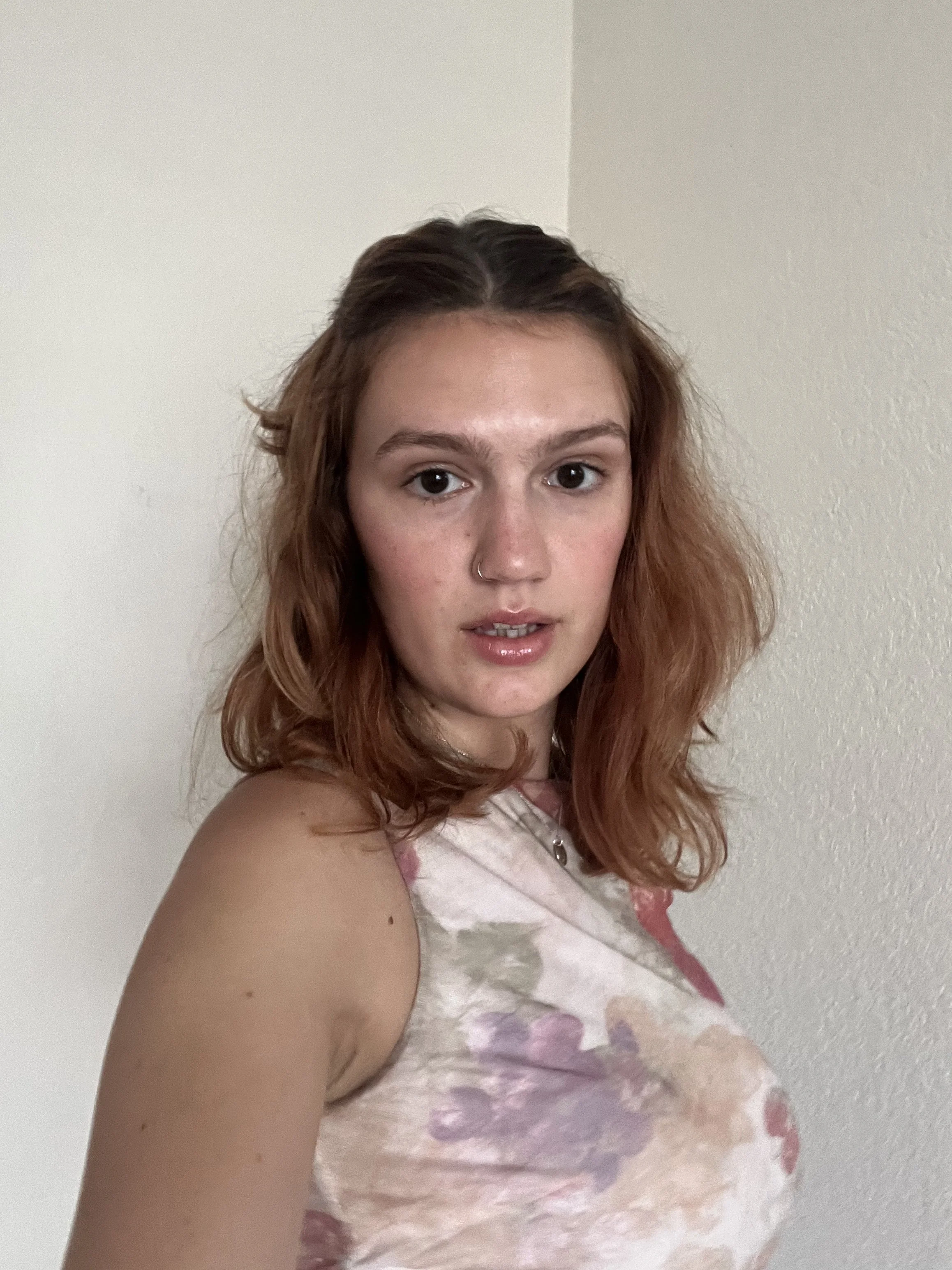 A young woman with shoulder-length curly red hair and a nose piercing, wearing a sleeveless floral top, standing indoors against a plain wall, looking at the camera.