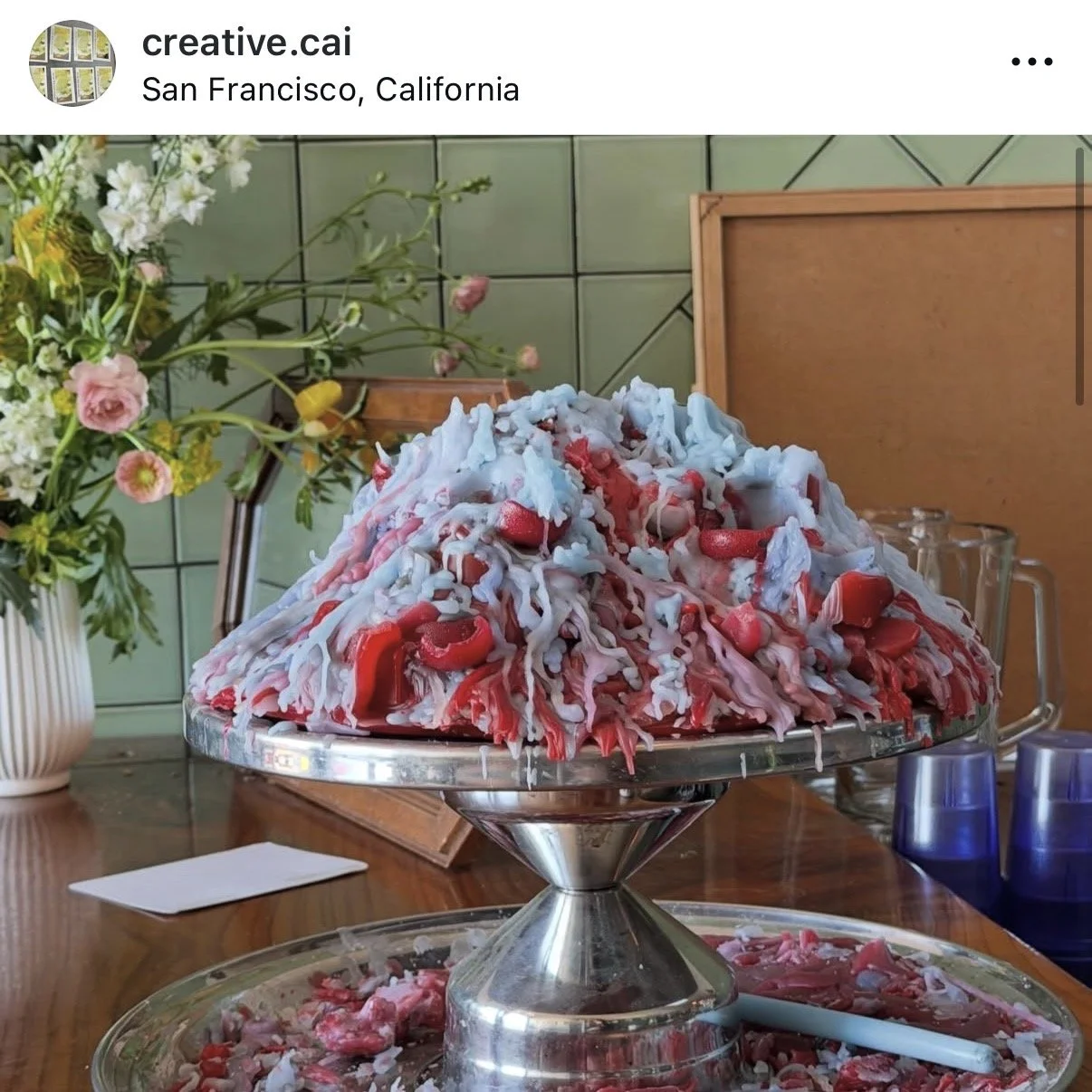 A large chocolate fountain with red and white melted candy flowing over a mound of candy on a stand, with glasses and flowers in the background.